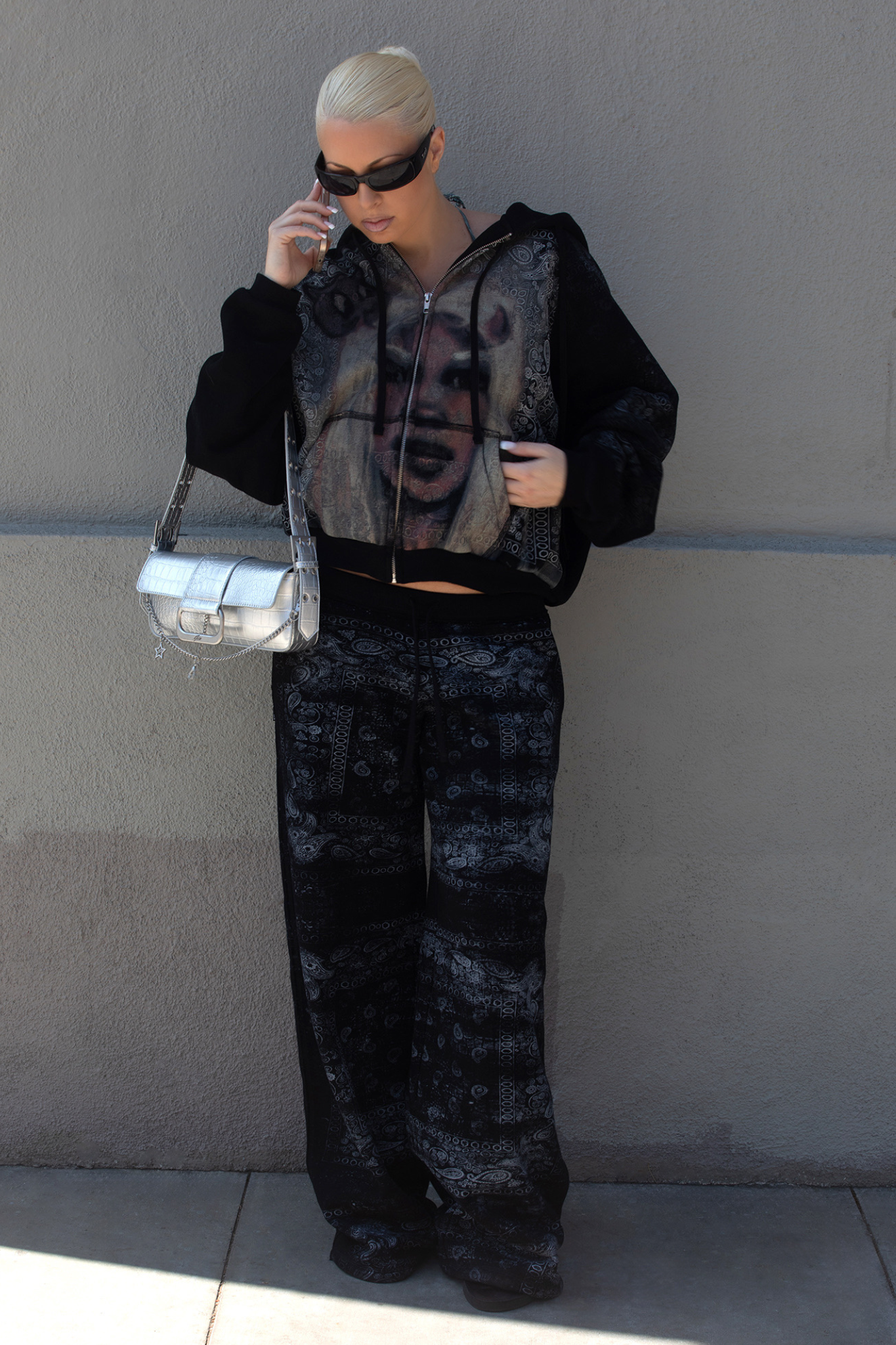 Wearing the I.AM.GIA T-REX ZIP THRU HOODIE in Black Bandana, a person with platinum blonde hair, sunglasses, and a silver purse pairs it with a dark patterned tracksuit and stands against a plain wall while talking on the phone.