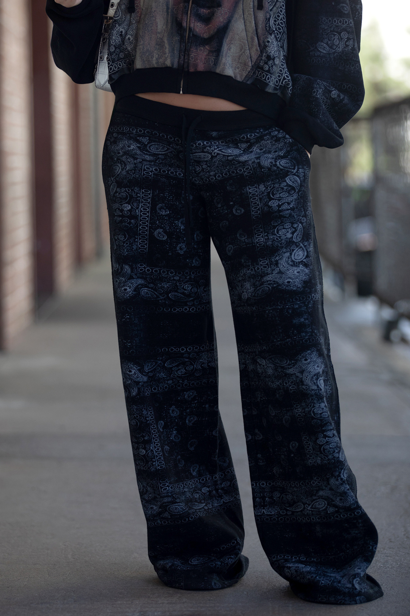 A person stands outdoors, showcasing the lower half of their outfit in I.AM.GIA’s T-REX TRACKPANT in Black Bandana, paired with a matching jacket; the street style aesthetic is highlighted against a softly blurred background.
