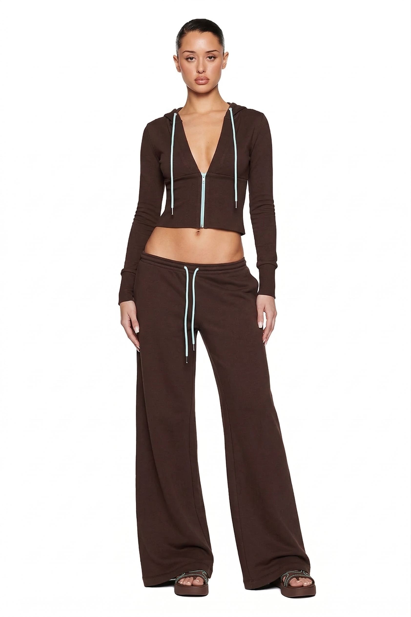 A woman poses against a white background wearing I.AM.GIA’s ARLO TRACK PANT in brown and blue, styled with a matching chocolate brown French terry zip-up crop hoodie, sandals, and her hair pulled back.