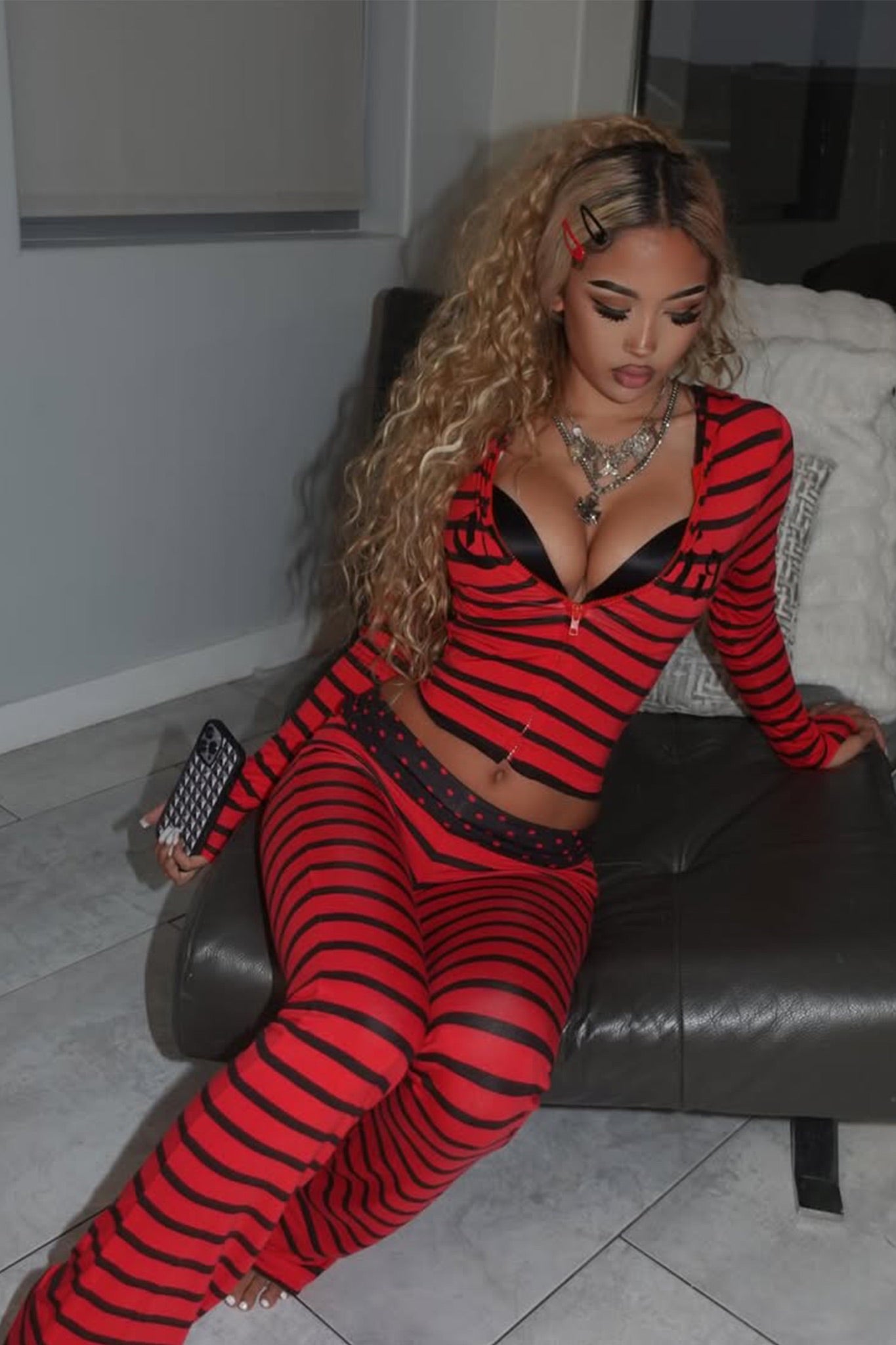 A woman with long curly blonde hair sits on a gray couch, wearing I.AM.GIA’s BLARE FOLDOVER TRACKPANT in red and black stripe and a black bra. She holds a phone, looking down, accessorized with hair clips and layered necklaces.