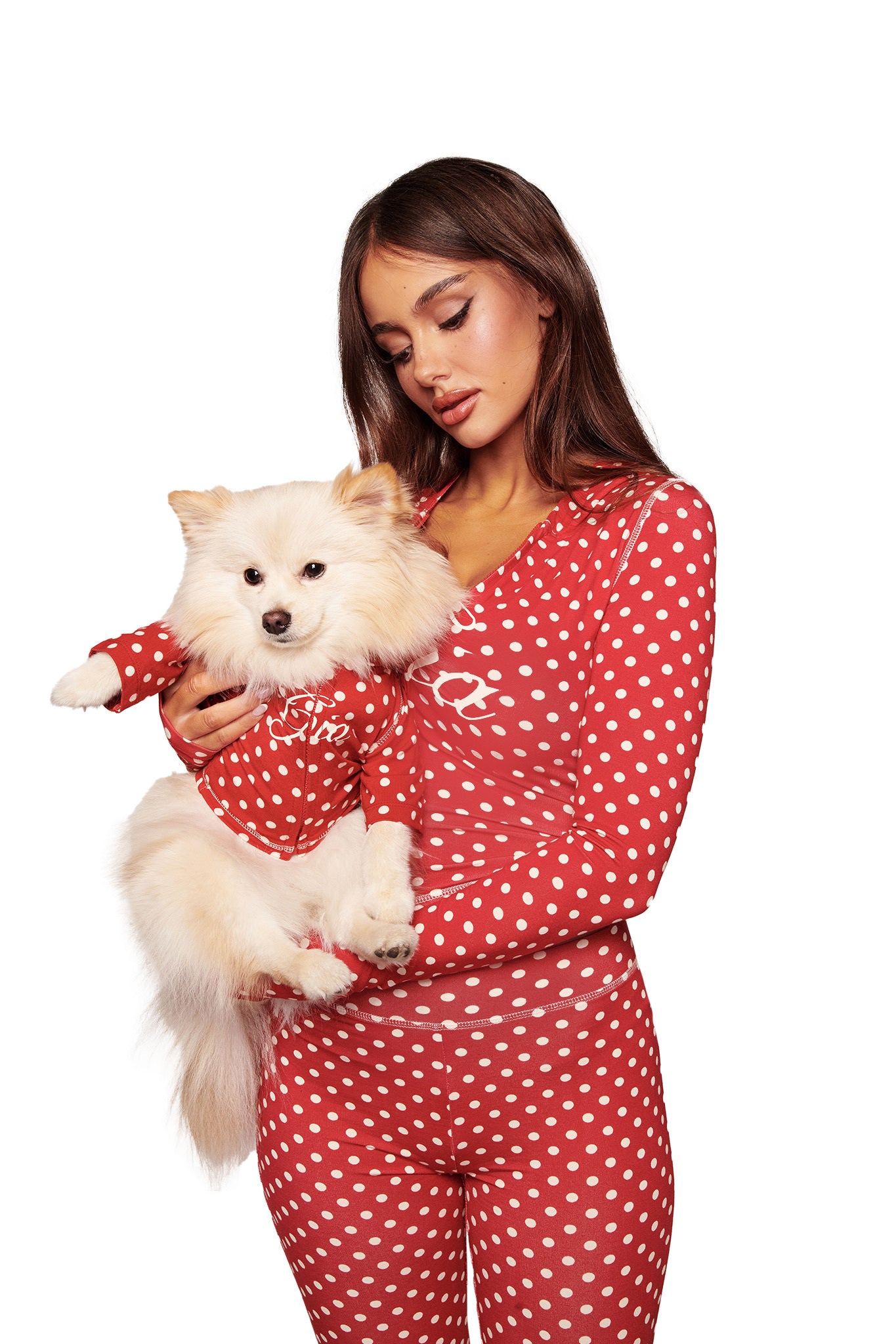 A woman in red-and-white polka dot pajamas holds a fluffy white dog wearing the I.AM.GIA BLARE BEST FRIEND DOG HOODIE - RED POLKA DOT. Both pose against a white background, showcasing their perfectly coordinated look.