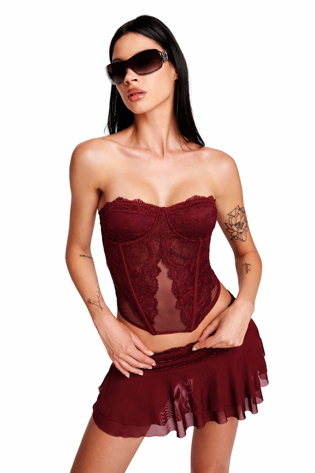 A woman with long dark hair and visible arm tattoos poses against a white background, wearing the I.AM.GIA DEMIE CORSET in deep red with sunglasses and a matching short ruffled skirt.