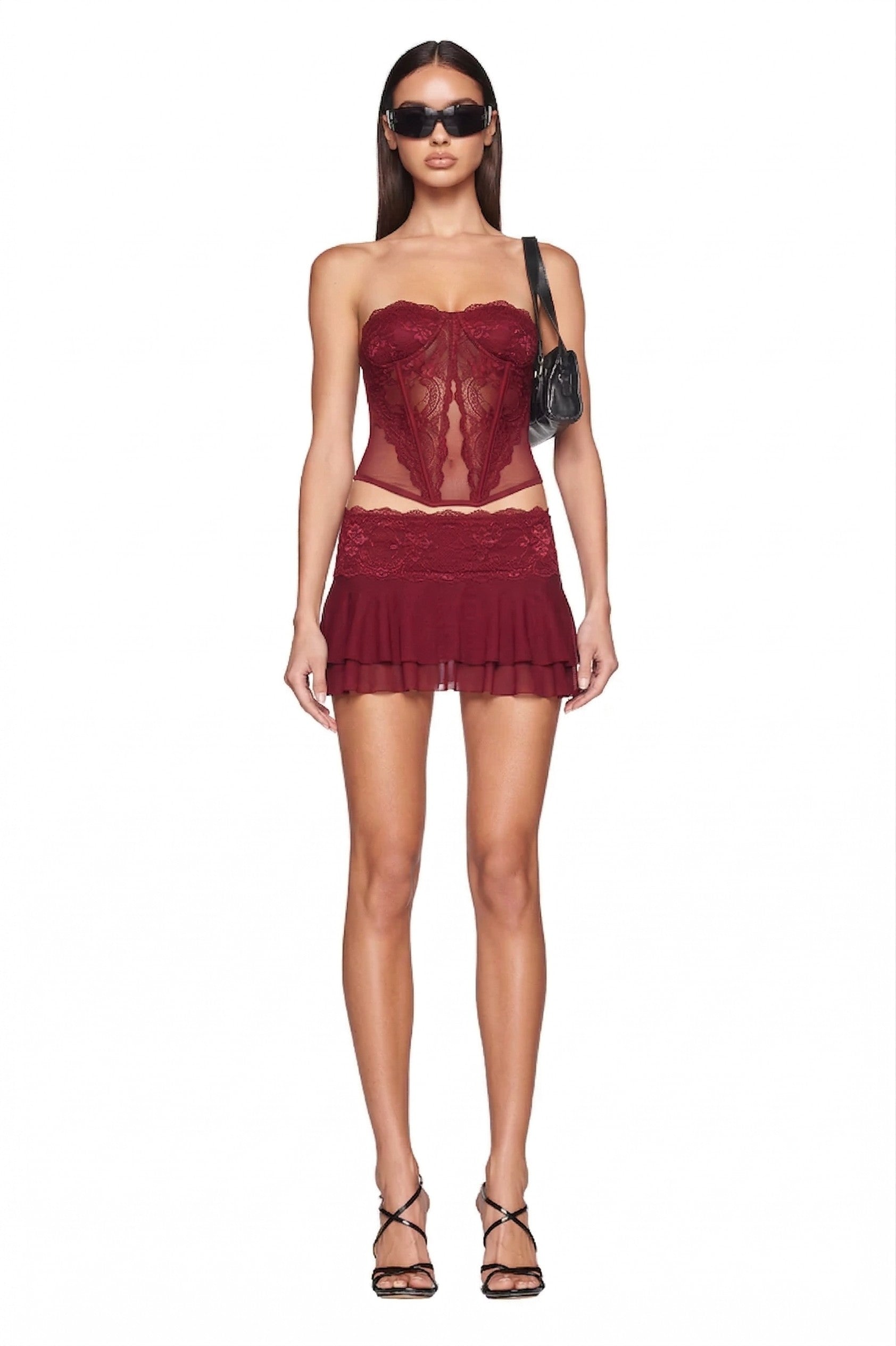 A woman wearing the I.AM.GIA DEMIE SKIRT in deep red with a strapless burgundy lace corset top, black strappy heels, sunglasses, and a small black shoulder bag stands against a white background.