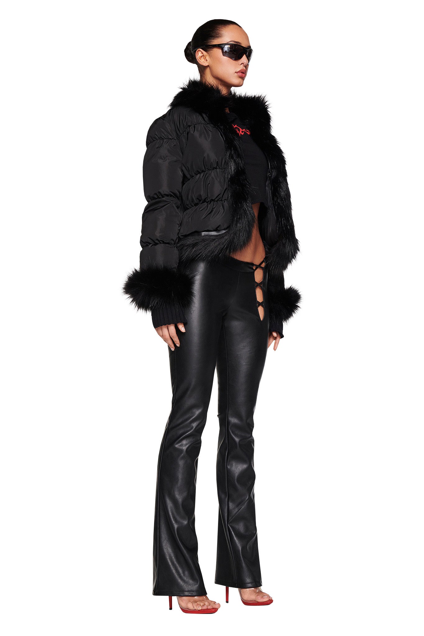 A person wearing black sunglasses, the BILLIE JACKET - BLACK from I.AM.GIA with fur trim, black leather pants featuring side cutouts, and red high-heeled shoes stands facing left. Their hair is tied back and they convey a stylish and confident appearance.