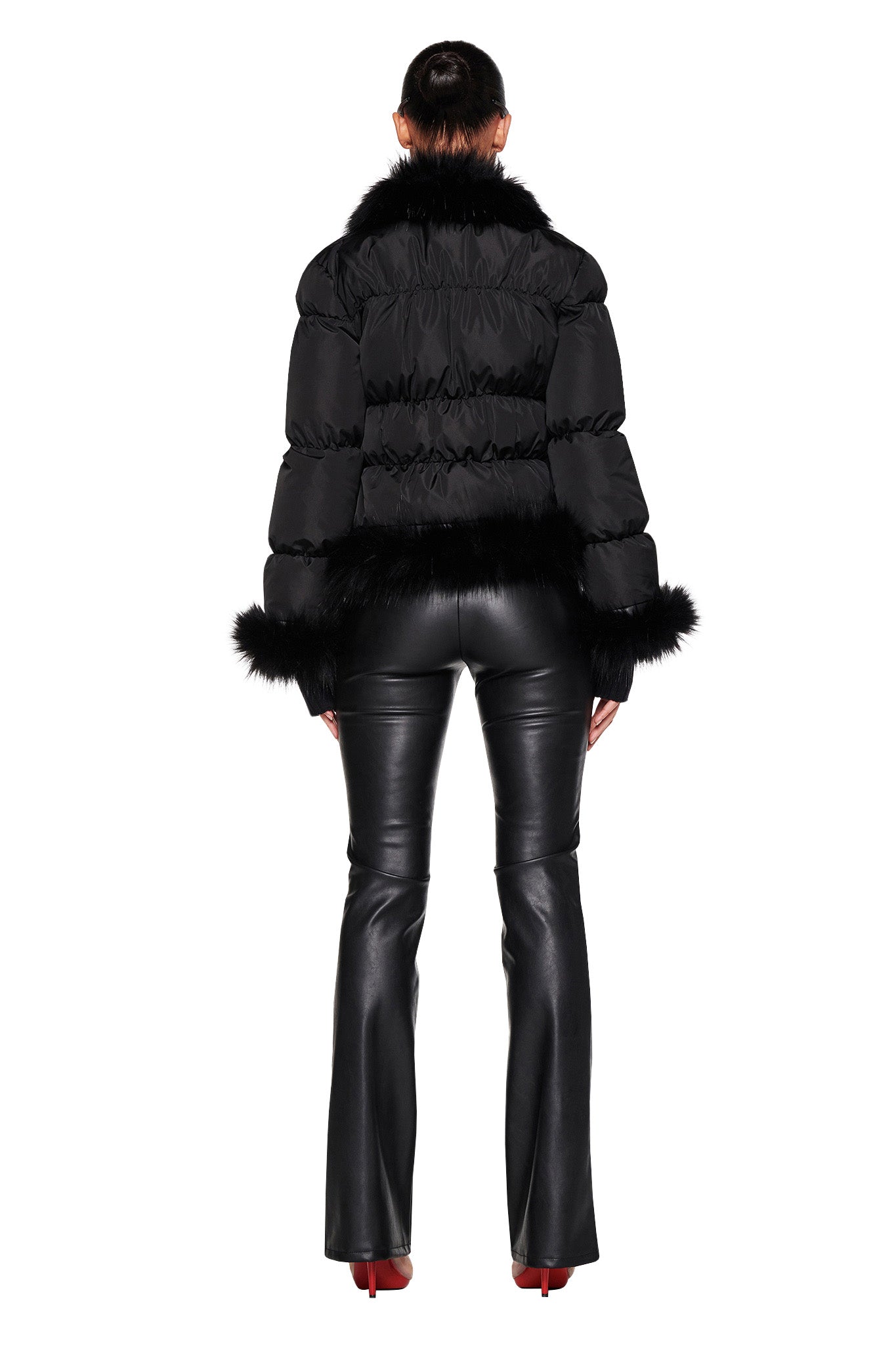 A person is facing away, wearing the BILLIE JACKET in black with fur trim on the collar and cuffs from I.AM.GIA. They are also sporting black, shiny leather pants and red high-heeled shoes. The background is white.