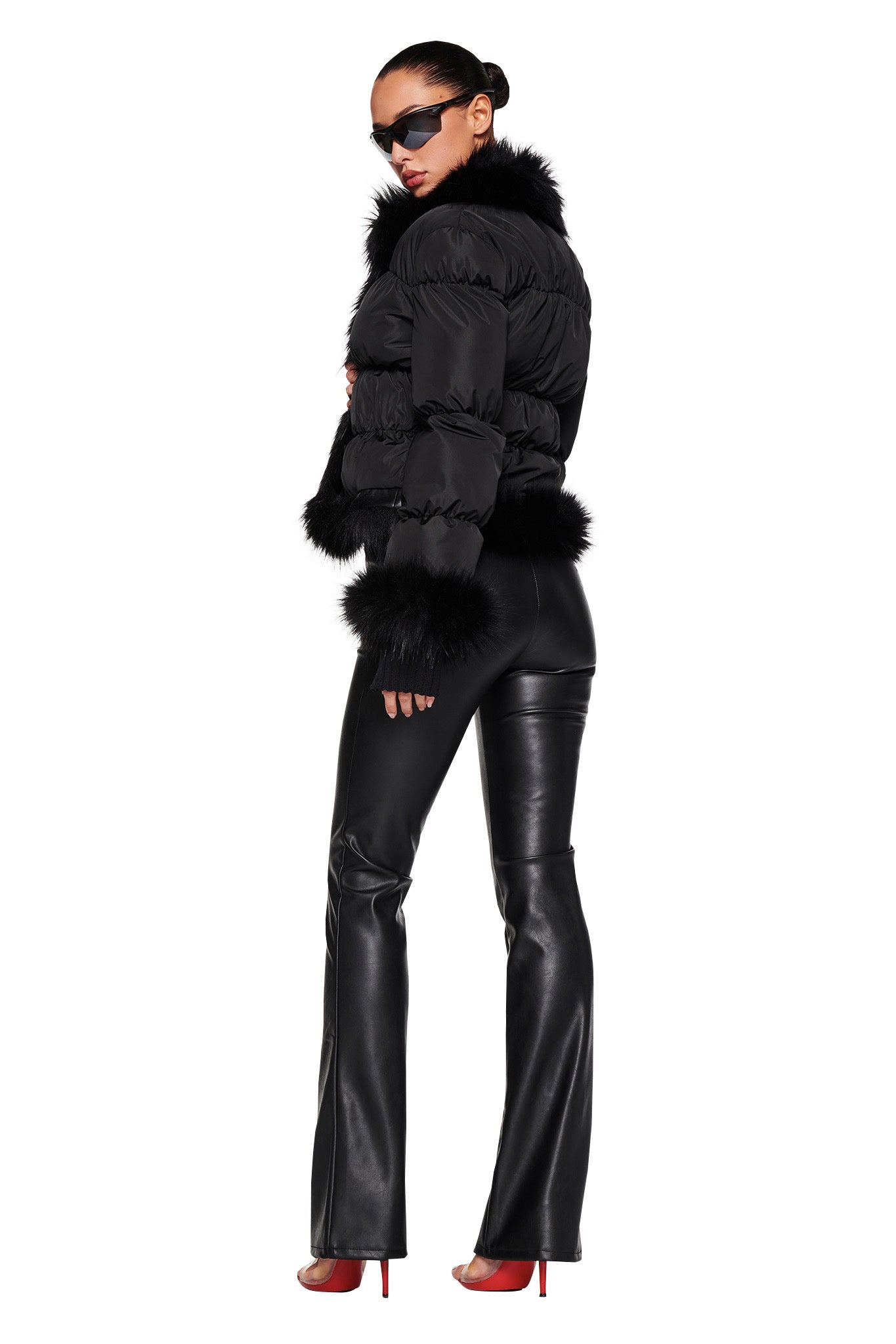 A woman stands confidently in an I.AM.GIA BILLIE JACKET - BLACK, fitted black leather pants, and red high heels. She accessorizes with dark sunglasses and has her hair tied back. The outfit exudes a chic, modern look with a touch of edginess.
