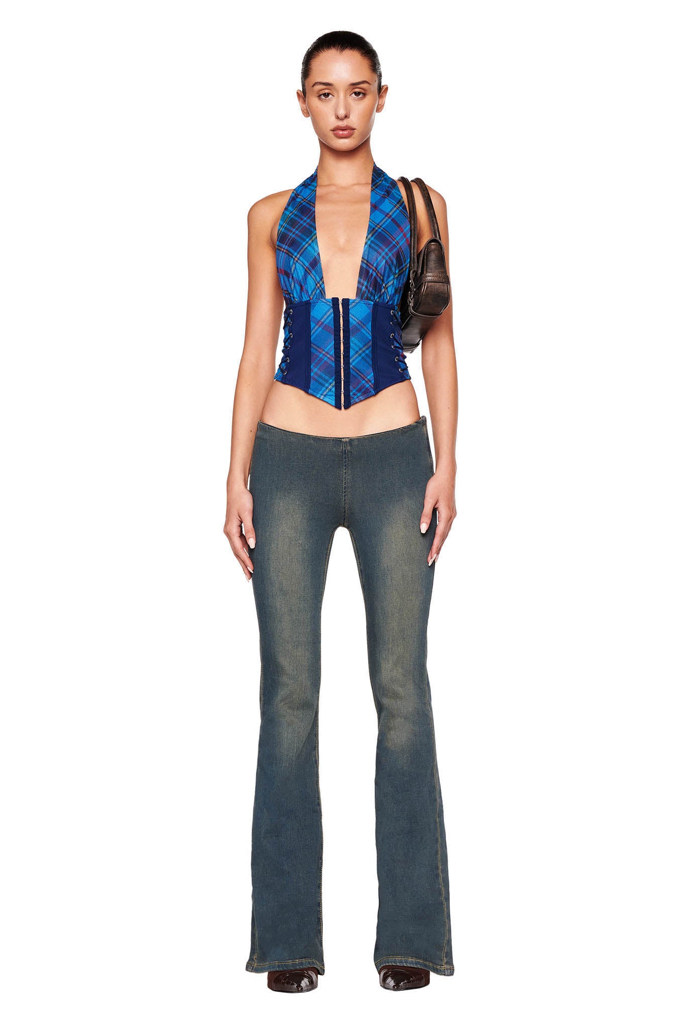 A person stands against a white background wearing the CELESTINA CORSET - BLUE : PLAID from I.AM.GIA, paired with low-rise bell-bottom jeans, and holding a small dark brown shoulder bag. The outfit is stylishly modern and form-fitting.