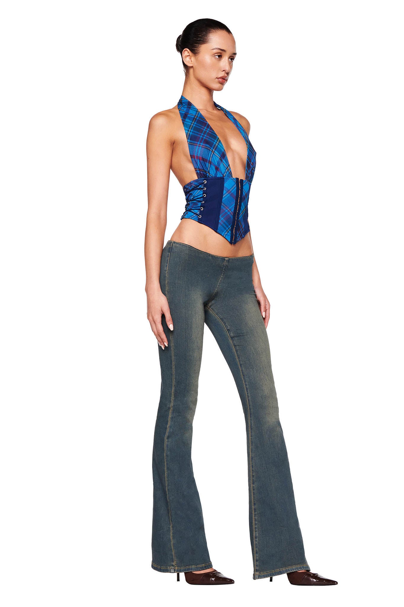 A person with a sleek bun hairstyle is modeling an I.AM.GIA fashion outfit featuring the CELESTINA CORSET - BLUE : PLAID, a blue plaid sleeveless top with a deep V-neck, paired with high-waisted, flared blue jeans. They complete the look with brown pointed-toe shoes against a pure white background.