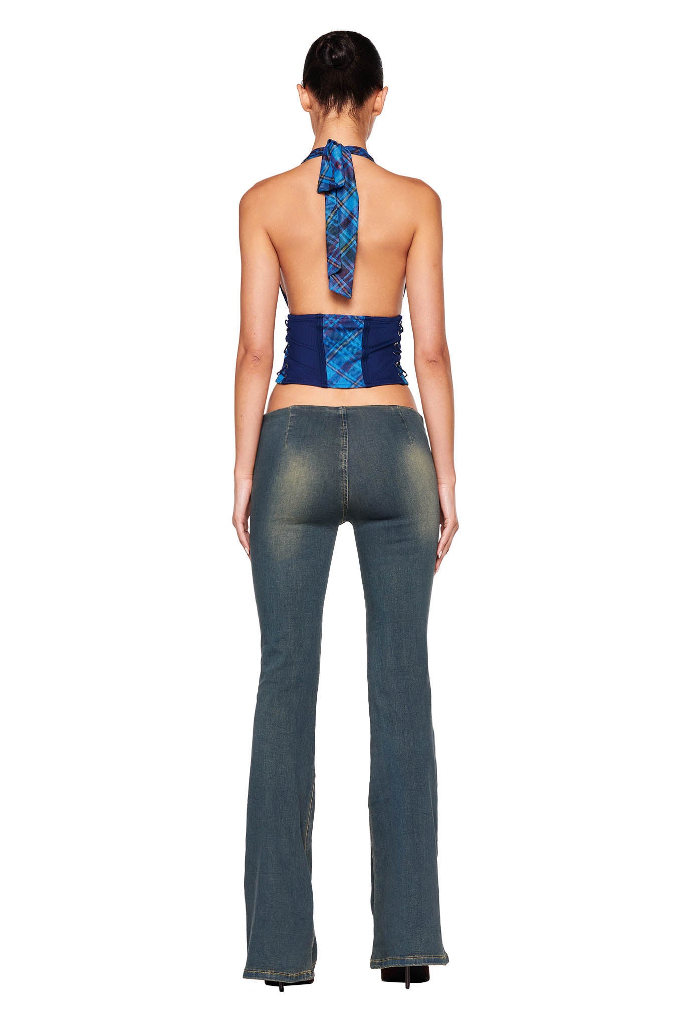A person is standing with their back to the camera, showcasing the details of their outfit. They are wearing the I.AM.GIA CELESTINA CORSET – BLUE : PLAID, which is a strapless top featuring a large plaid bow tied at the neck that complements their bare back. The look is paired with faded blue bell-bottom jeans for a chic finish.