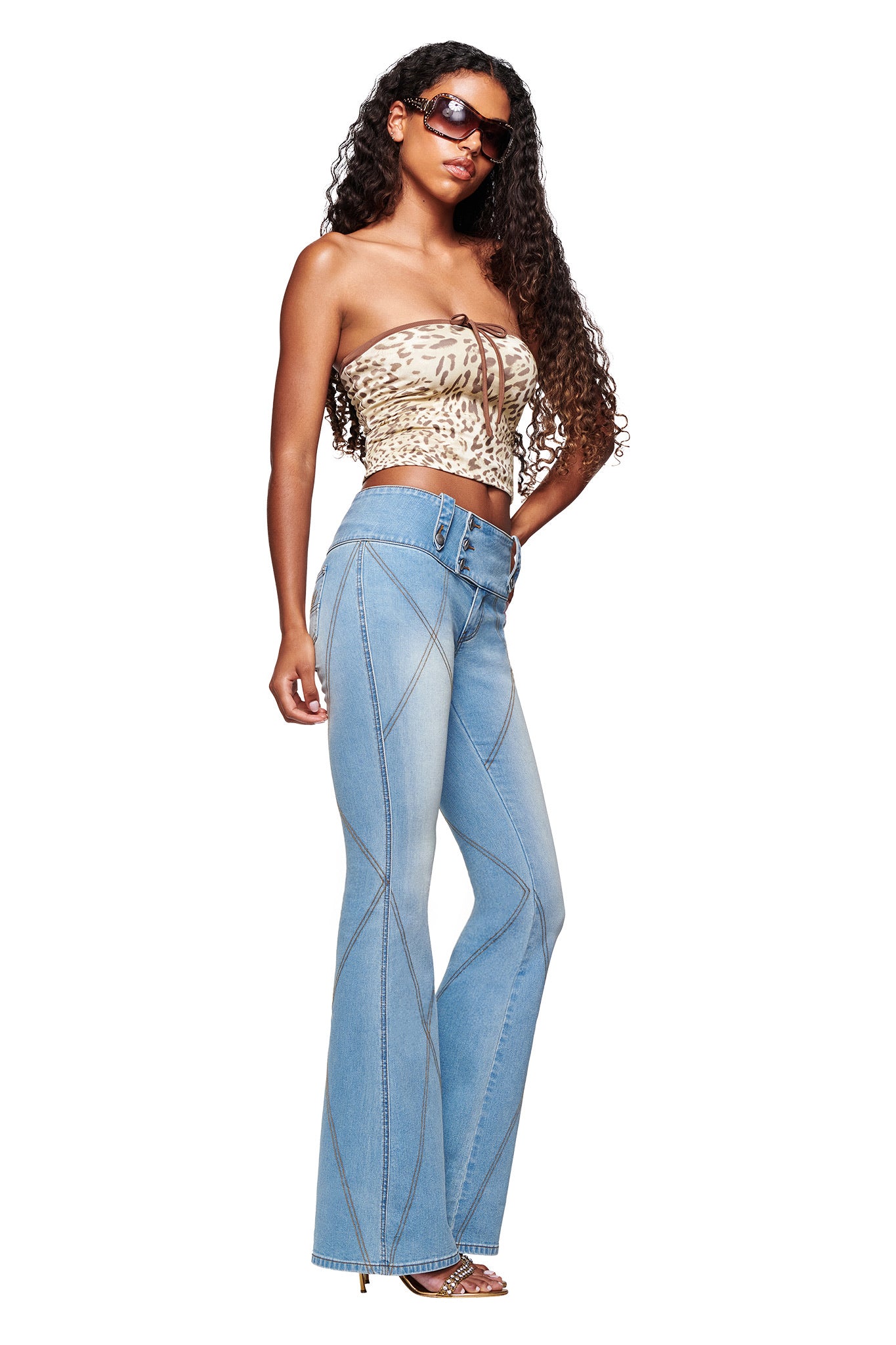 A woman with long curly hair wears sunglasses, a strapless leopard print top, I.AM.GIA’s MALLORY JEAN - LIGHT BLUE DENIM flared jeans, high heels, and poses against a white background.