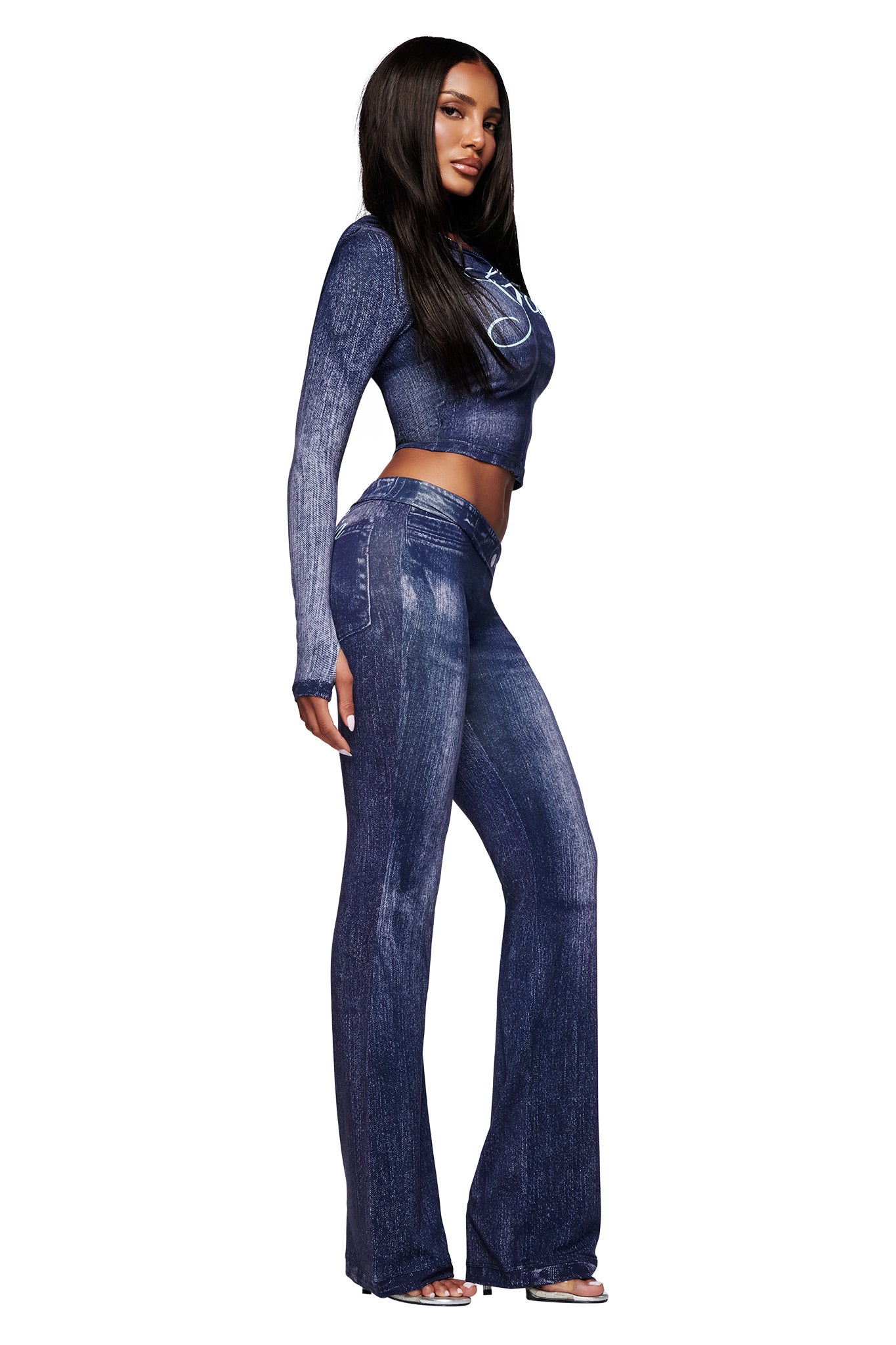 A woman stands in profile against a white background, wearing I.AM.GIA’s BLARE TRACKPANT - DARK DENIM and a matching blue, long-sleeve crop top. She has long straight hair and open-toed shoes.
