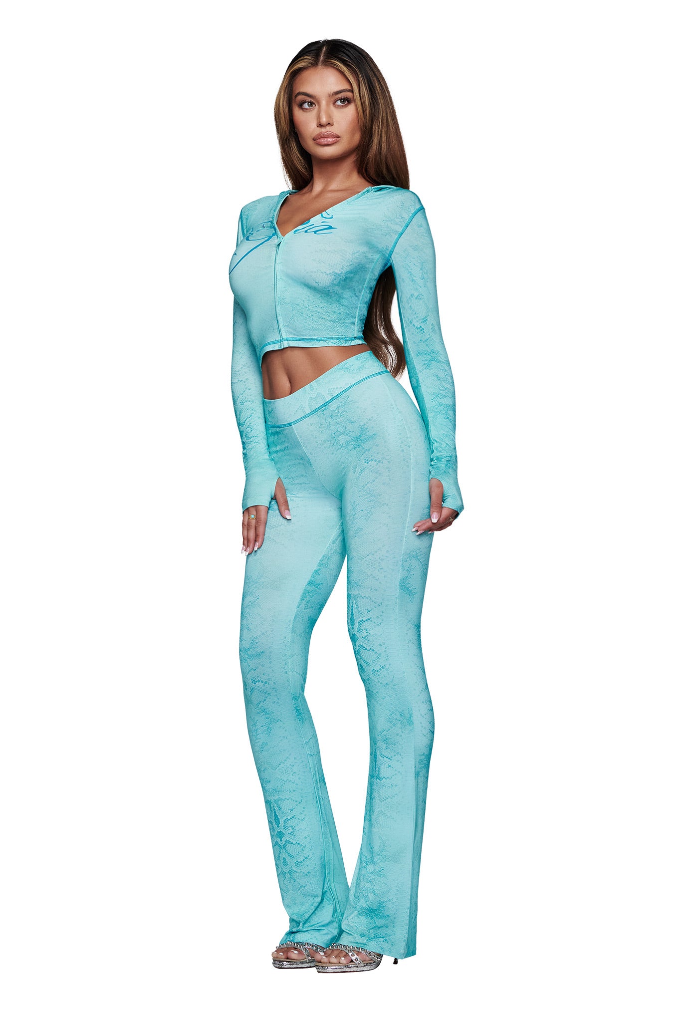 A woman stands against a white background wearing the I.AM.GIA BLARE HOODIE in Teal Serpentine, styled with long, straight hair and clear high-heeled shoes.