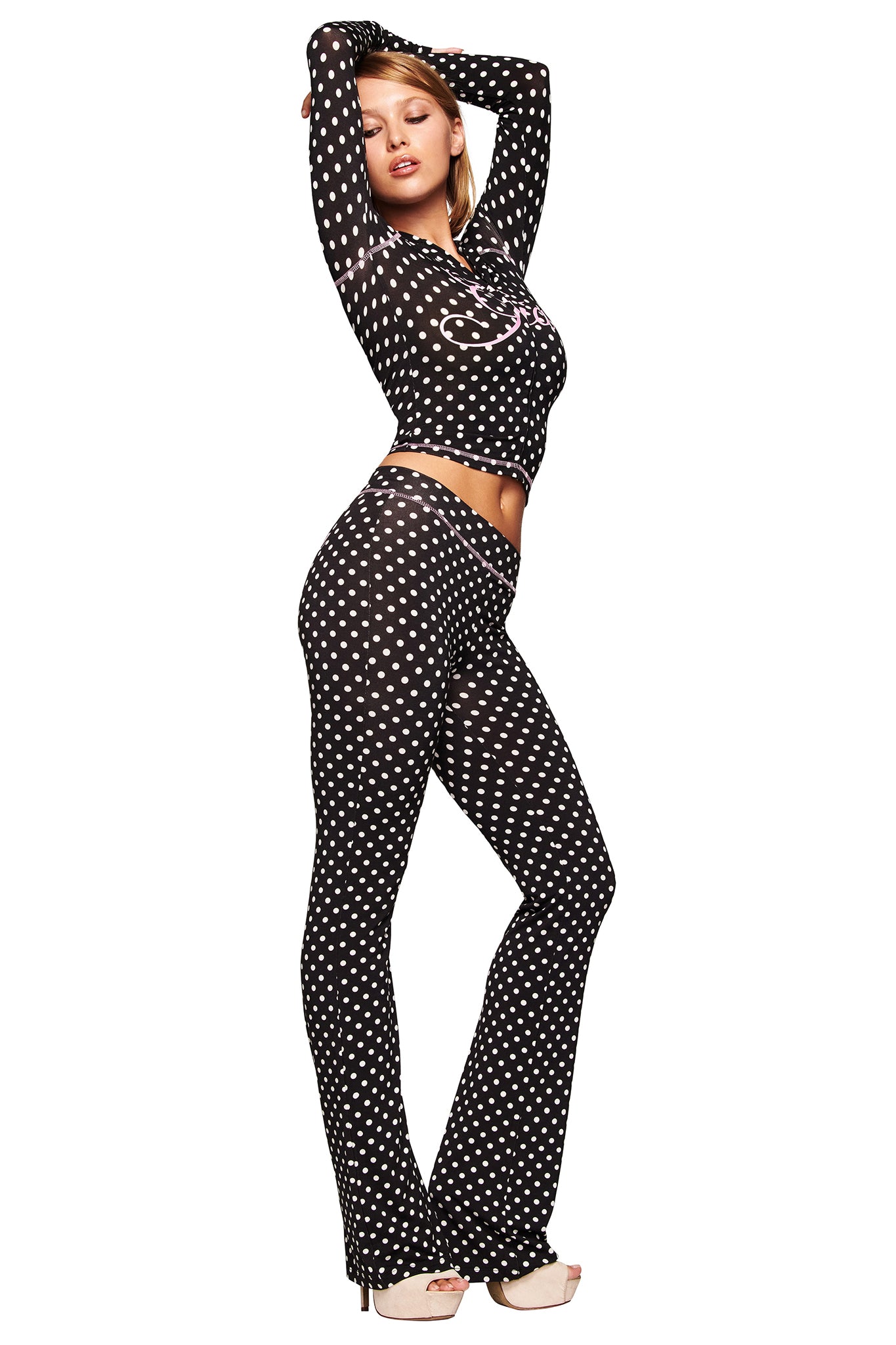 A woman poses in the I.AM.GIA BLARE HOODIE - BLACK POLKA DOT with matching flared pants and beige heels, one arm raised above her head against a white background.