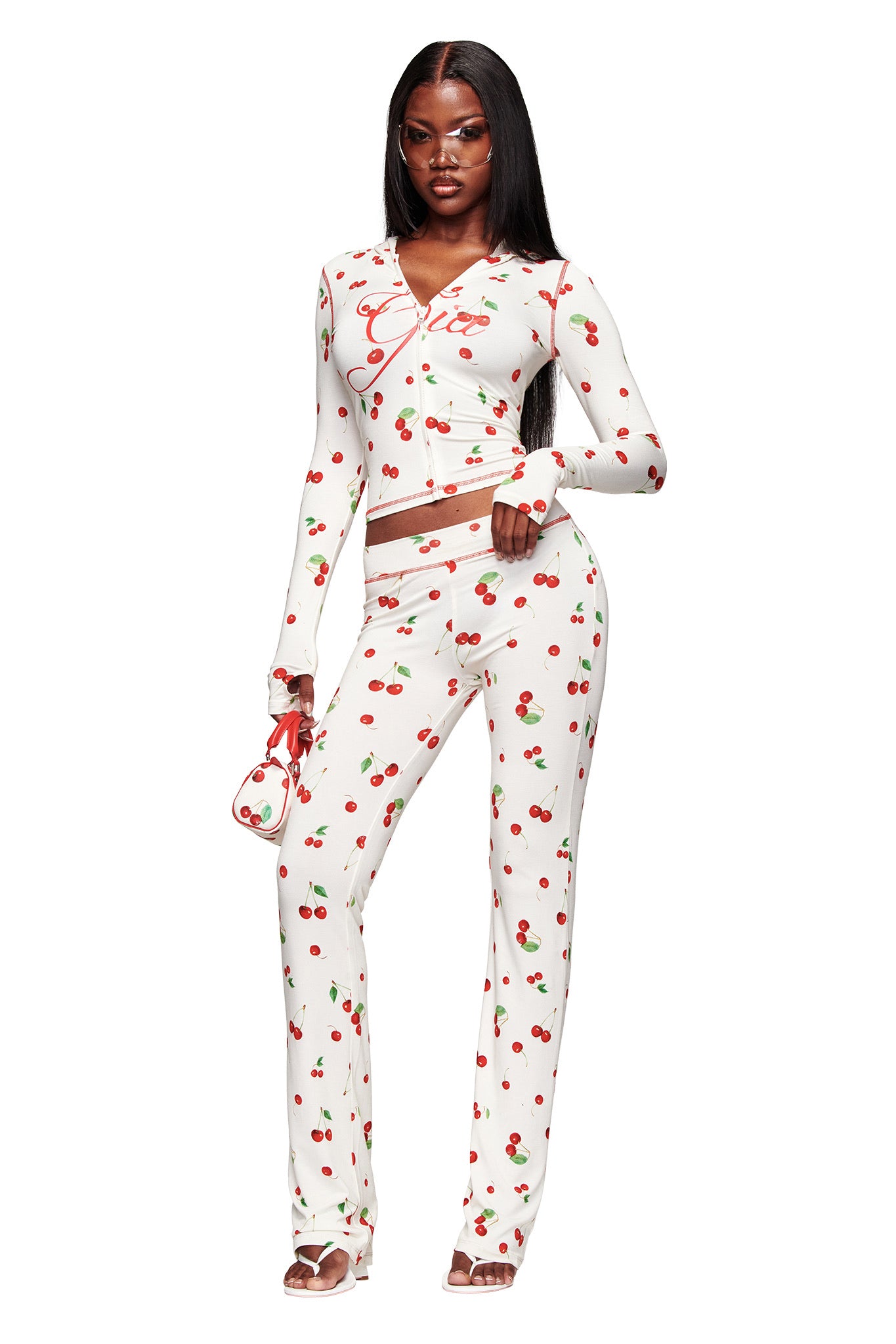 A woman wears the I.AM.GIA BLARE TRACKPANT - CHERRY ON TOP, styled as a two-piece with long sleeves and a matching purse. She poses in heeled sandals against a white background.