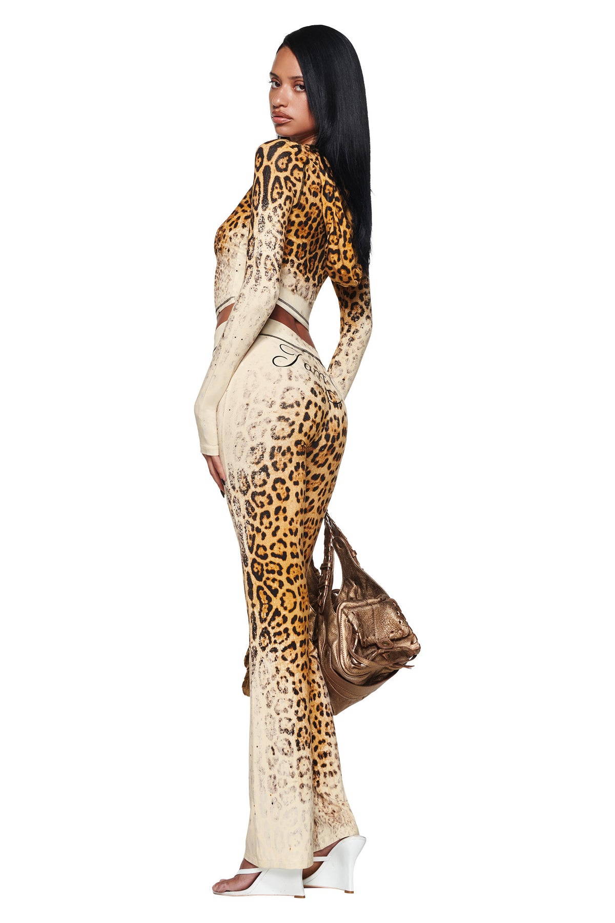 A person in a cream and leopard print BLARE HOODIE - CREAM FADED PANTHERA and matching flared pants stands in profile, holding a metallic bronze handbag.