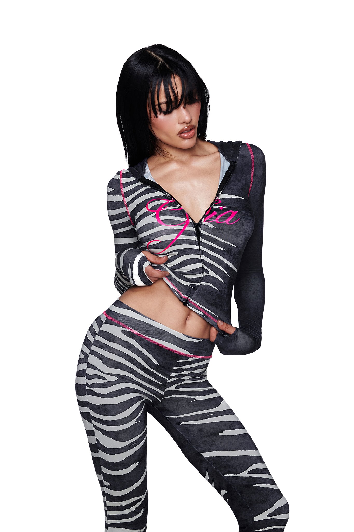 A woman poses against a white background in the BLARE TRACKPANT - BLACK WILD SIDE by I.AM.GIA, featuring a striking zebra print and matching zip-up top. With its mid-rise waist that offers a flattering fit and crafted from breathable bamboo fabric for all-day comfort, the trackpant combines style and ease as she looks down, her hand lifting her shirt slightly.