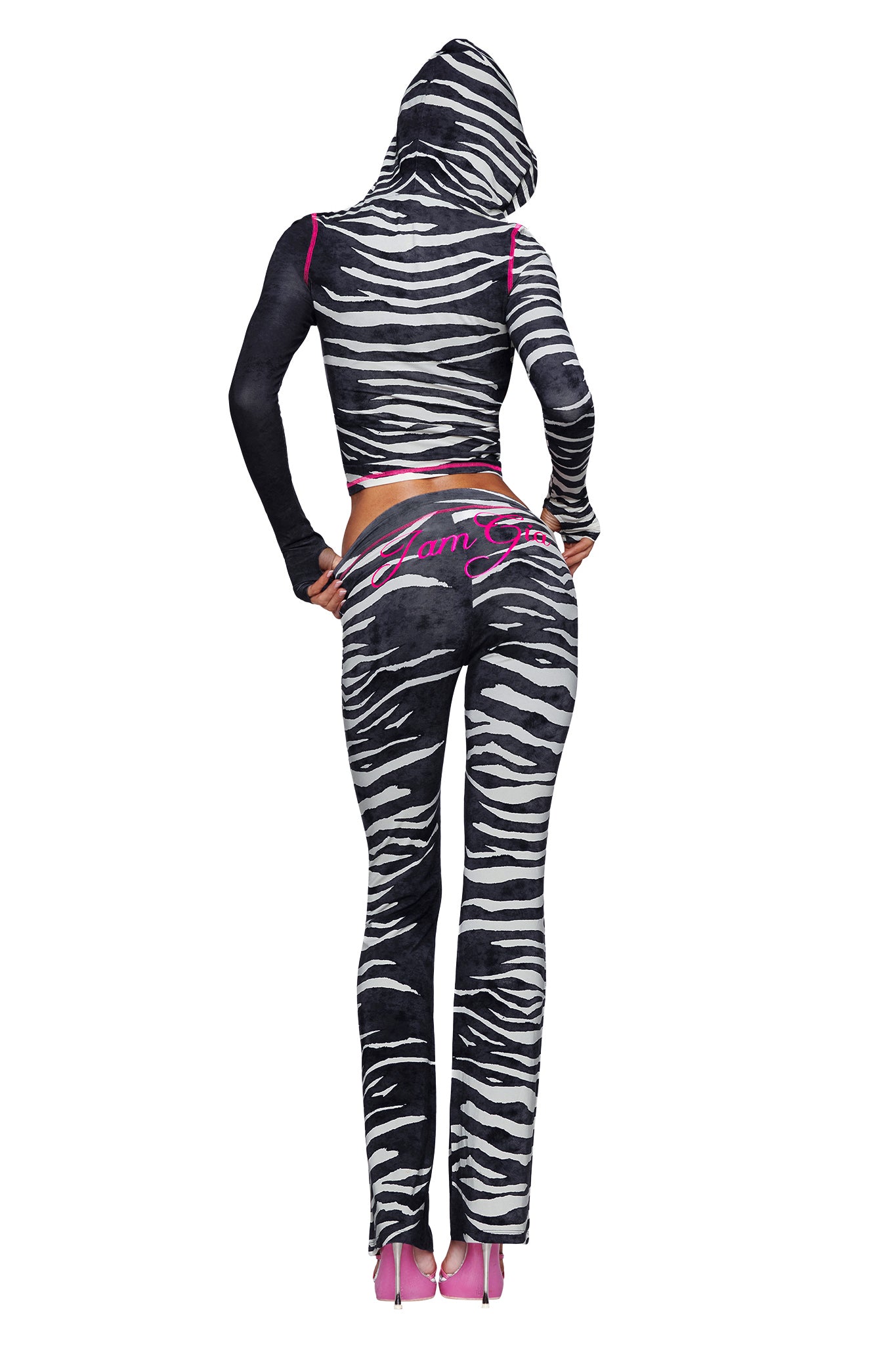 A person stands facing away, dressed in the I.AM.GIA BLARE HOODIE - BLACK WILD SIDE—a bold black-and-white zebra print hoodie—and matching pants with pink lettering on the back, paired with vibrant pink high heels.