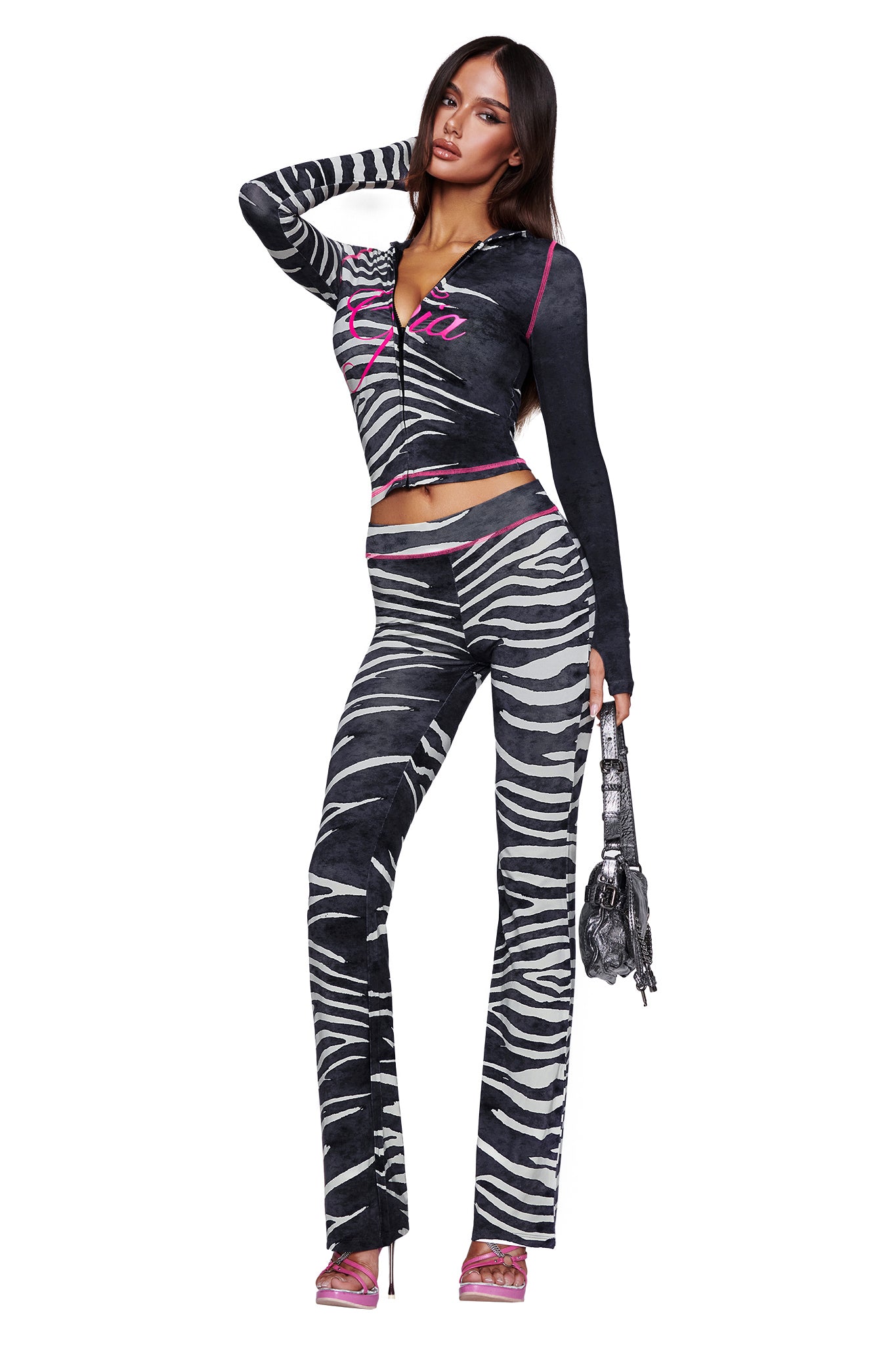 Wearing the I.AM.GIA BLARE TRACKPANT - BLACK WILD SIDE, a woman poses in a coordinated zebra-print set with flared pants and a zip-up top featuring a mid-rise waist. She completes the look with a small handbag and pink high heels.