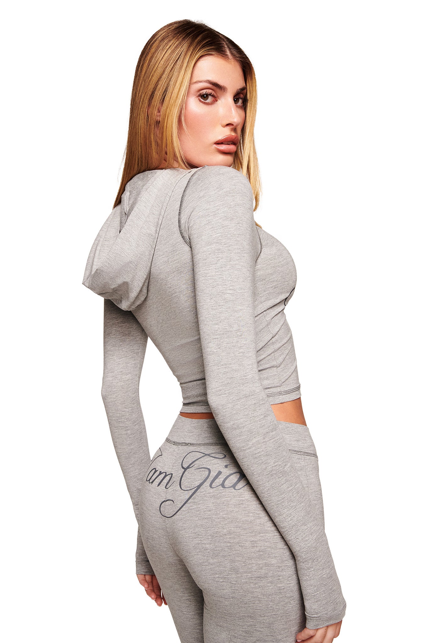 A woman models the I.AM.GIA BLARE JERSEY HOODIE in heather grey with matching high-waisted pants, her back turned to reveal "I am Gia" on the pants, set against a plain white background.