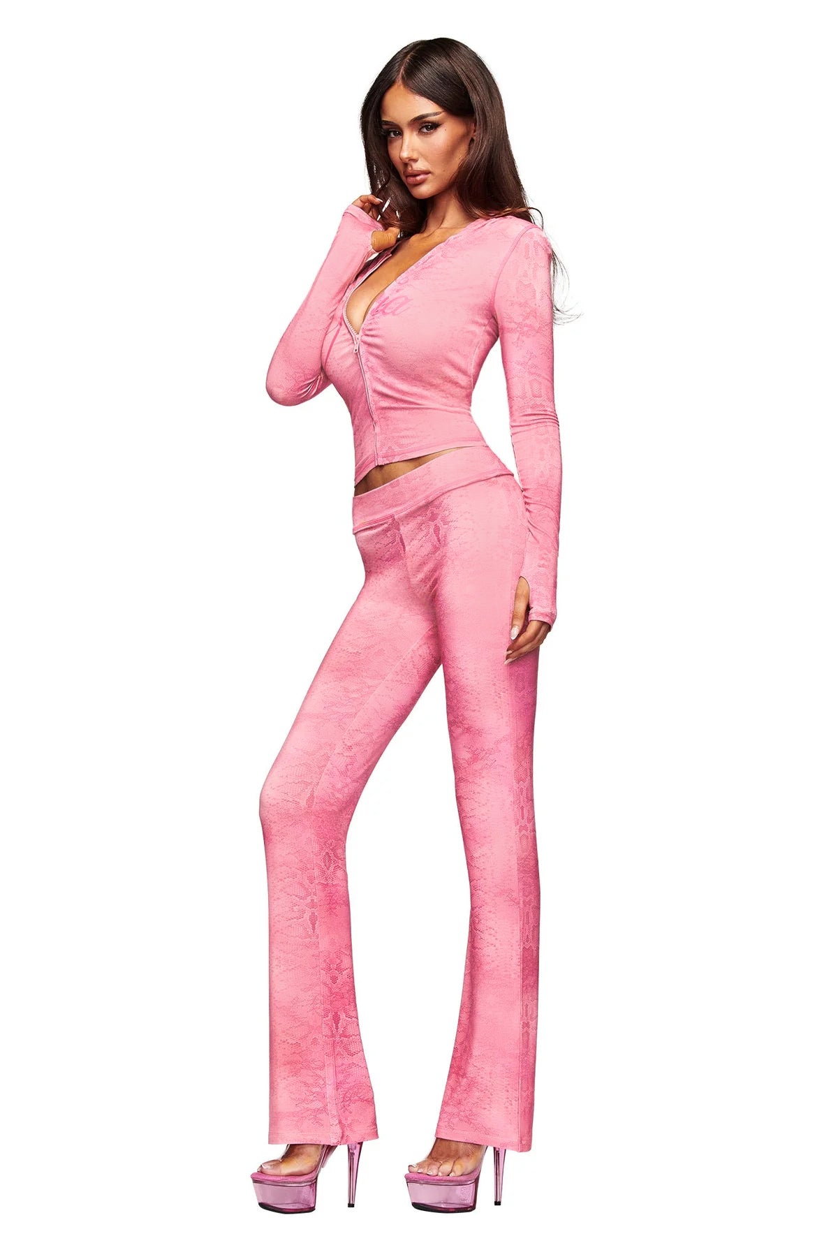 A woman poses confidently against a white background wearing the I.AM.GIA BLARE TRACKPANT - LIGHT PINK SERPENTINE, paired with a fitted, light pink long-sleeve top and pink platform heels.