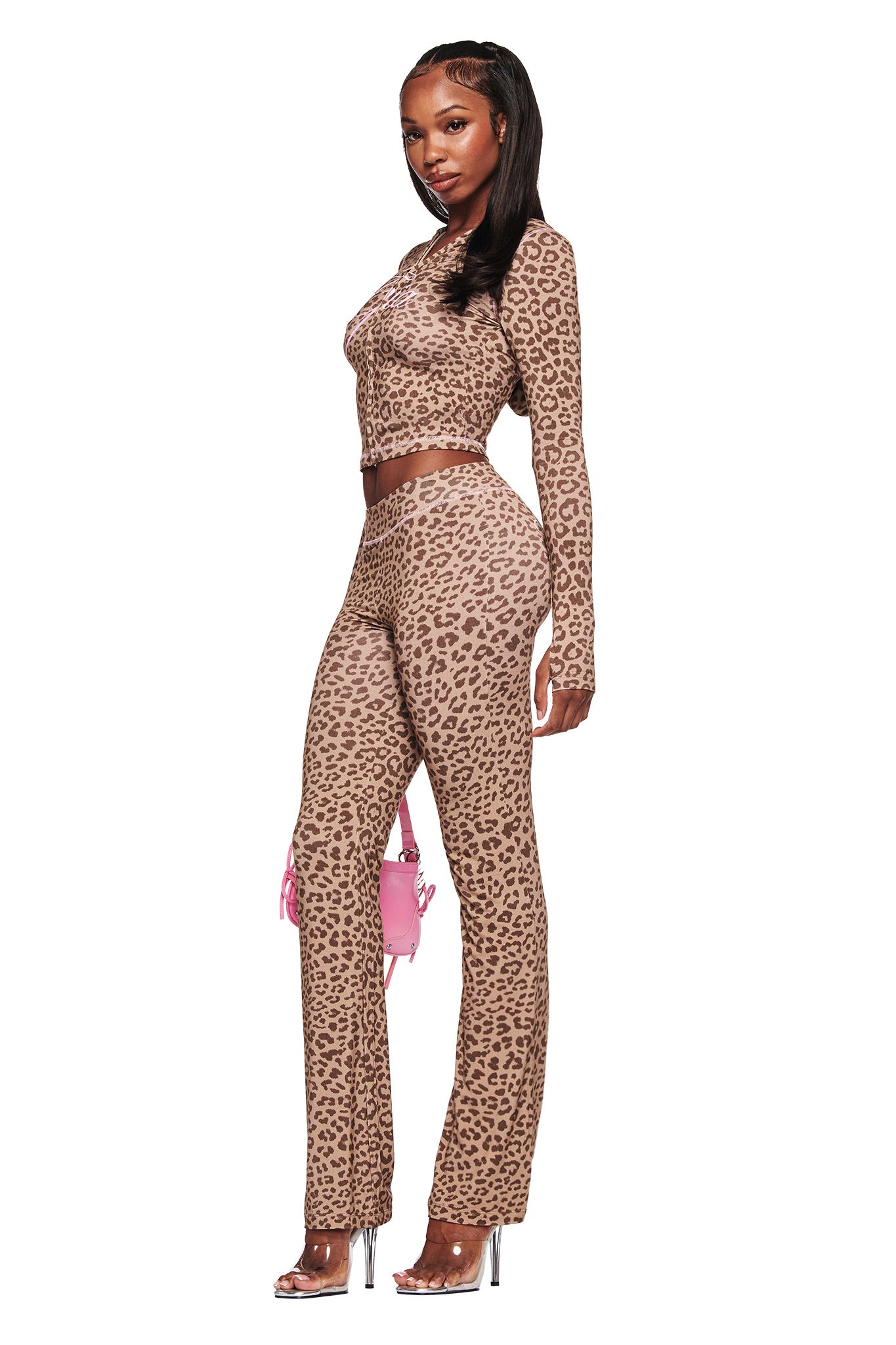 A woman in profile wears the I.AM.GIA BLARE TRACKPANT - MOCHA LEOPARD and matching crop top, styled with clear high heels and a small pink handbag, set against a white background.