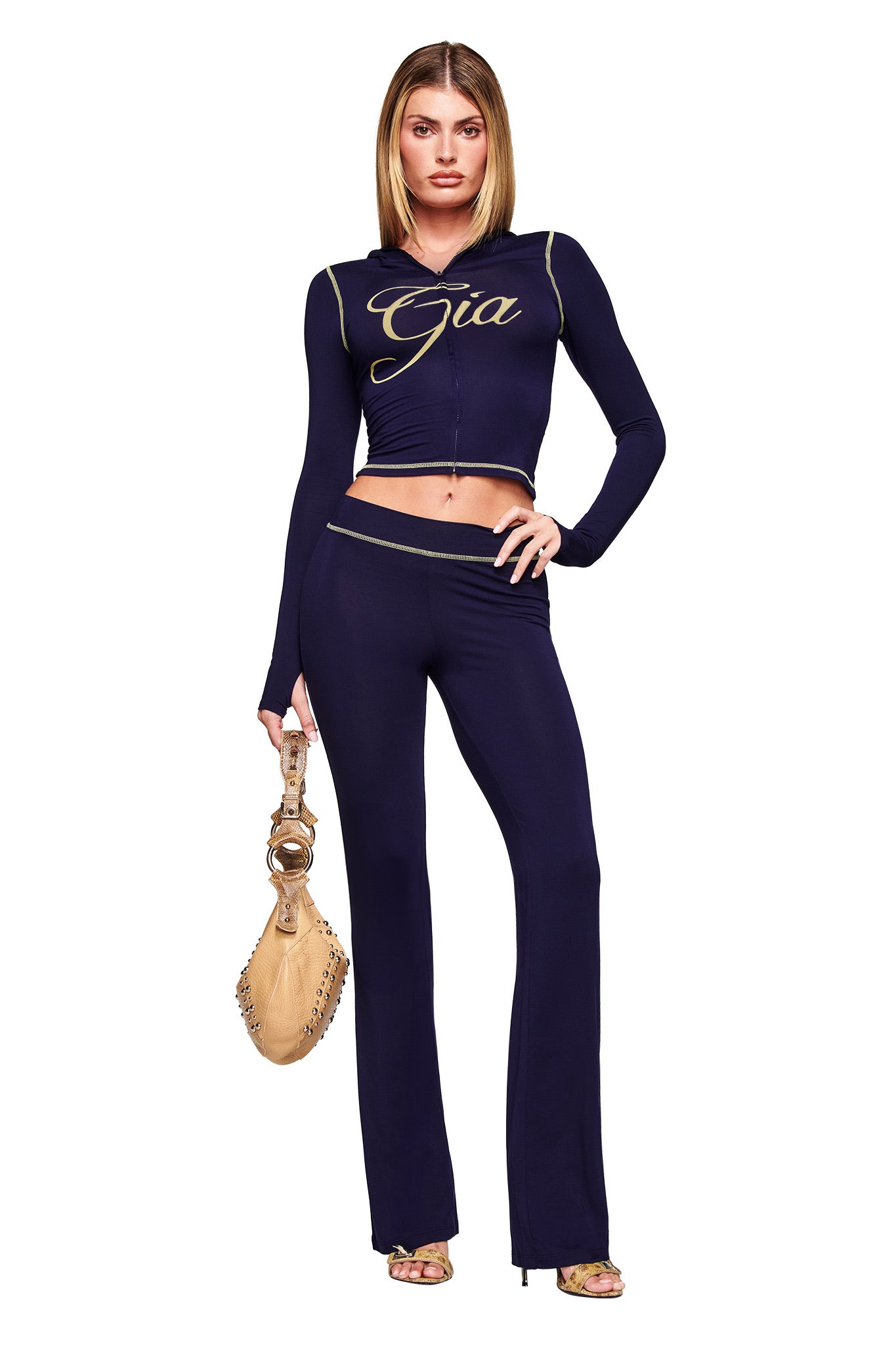 A woman stands confidently in I.AM.GIA’s navy BLARE TRACKPANT, a navy long-sleeve cropped top with “Gia” in gold, gold sandals, and a tan handbag against a plain white background.