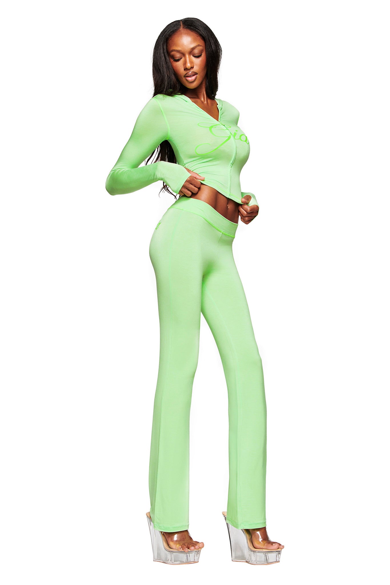 A woman models the I.AM.GIA BLARE TRACKPANT in neon green, featuring a low-rise waist, flared legs, and a matching crop top with cutouts. She completes the bold look with clear platform heels against a white background.