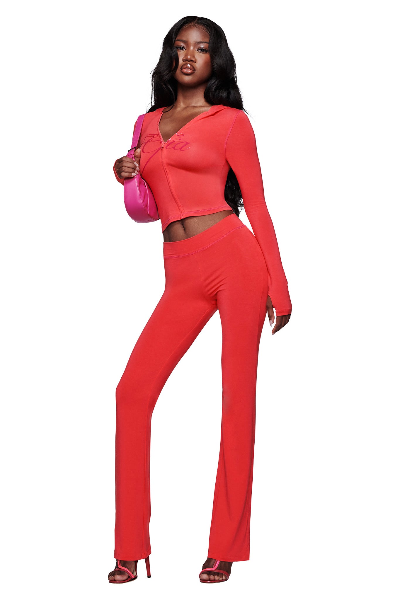 A woman with long, wavy black hair models I.AM.GIA's BLARE TRACKPANT - PAPAYA, a fitted flared leg pant paired with a matching long-sleeve top, red heeled sandals, and a pink shoulder bag against a white background.