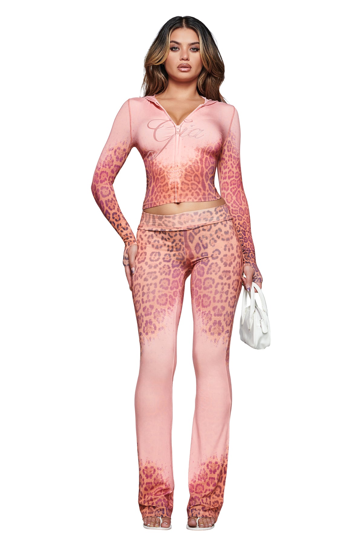 A woman stands against a white background wearing the I.AM.GIA BLARE HOODIE in Pink Faded Panthera print, styled with matching flared pants, a small white handbag, and clear high-heeled shoes.