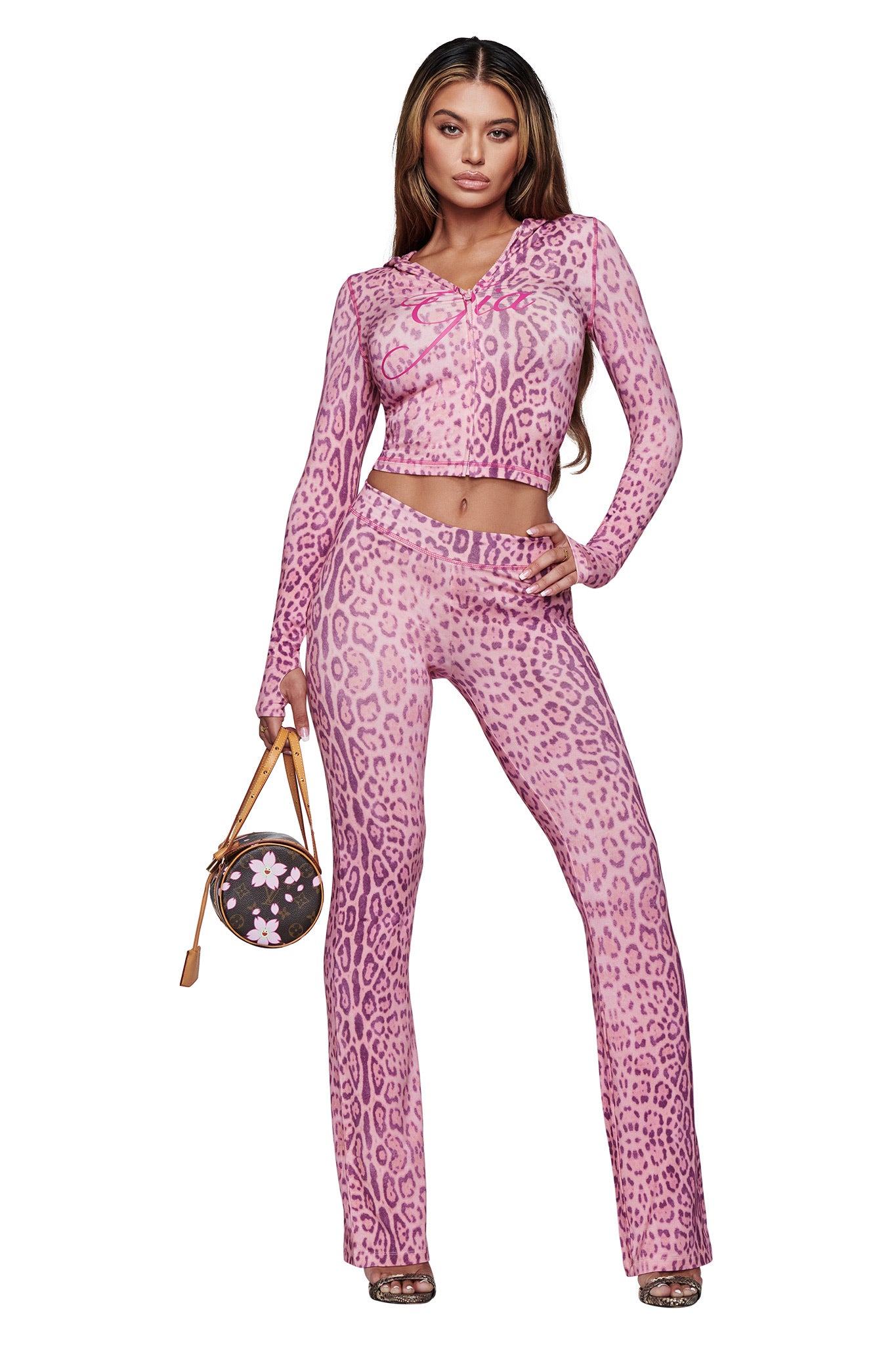 A woman poses confidently in the I.AM.GIA BLARE TRACKPANT - PINK PANTHERA set, paired with a round brown handbag adorned with pink florals and high-heeled sandals.