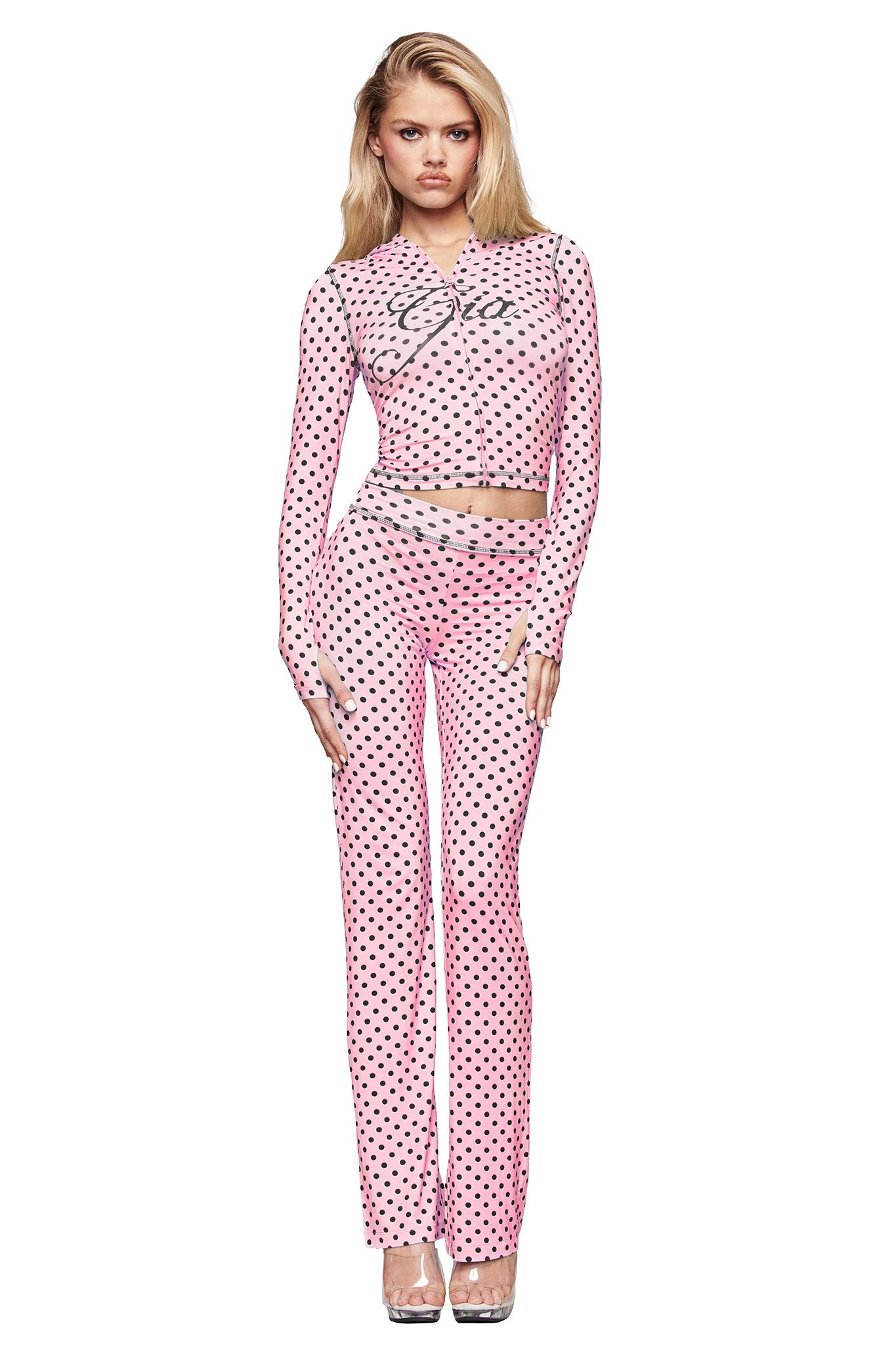 A woman with long blonde hair faces forward, wearing I.AM.GIA’s BLARE TRACKPANT in pink polka dot with a matching fitted, long-sleeve mesh top and clear high-heeled shoes, posed against a white background.