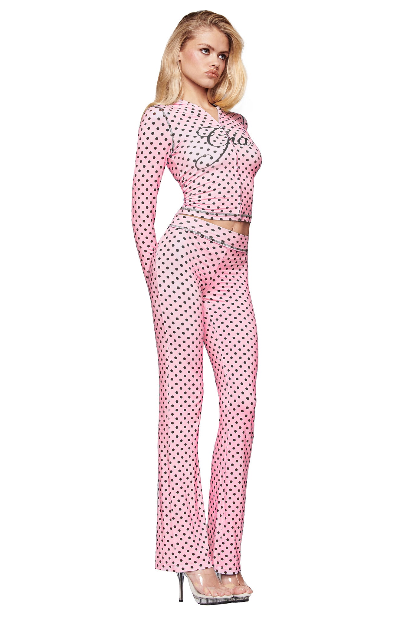 A woman in profile wears a fitted, long-sleeve pink top with decorative writing and I.AM.GIA BLARE TRACKPANT - PINK POLKA DOT, paired with silver heels and her blonde hair down.