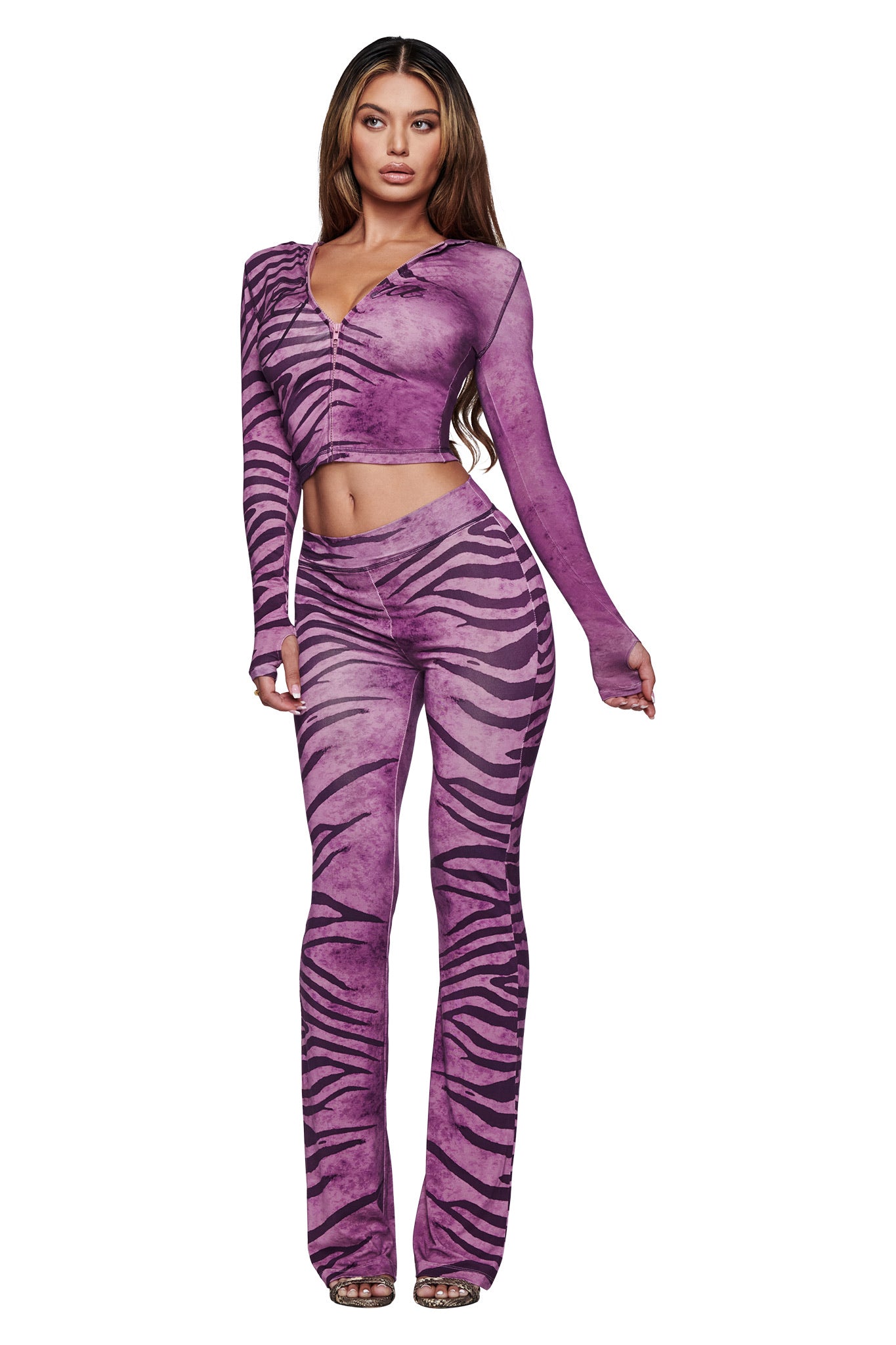 A woman stands against a white background wearing the I.AM.GIA BLARE TRACKPANT - PLUM WILD SIDE, a high-waisted, flared pant with a zebra print, paired with a matching fitted long-sleeve crop top.