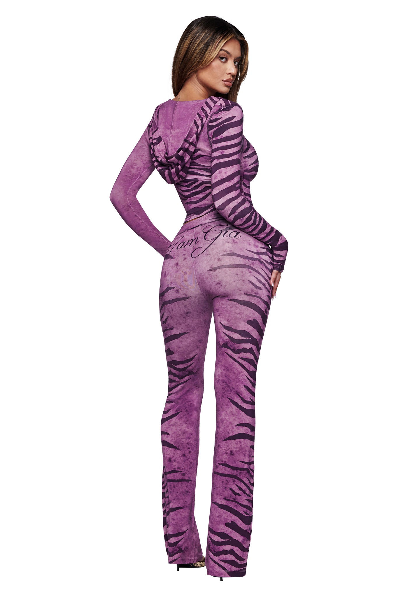 A woman faces away wearing the I.AM.GIA BLARE TRACKPANT in Plum Wild Side, featuring a tiger-striped design, flared legs, and full-length coverage. She turns her head to the side, showing her profile with one hand on her hip.