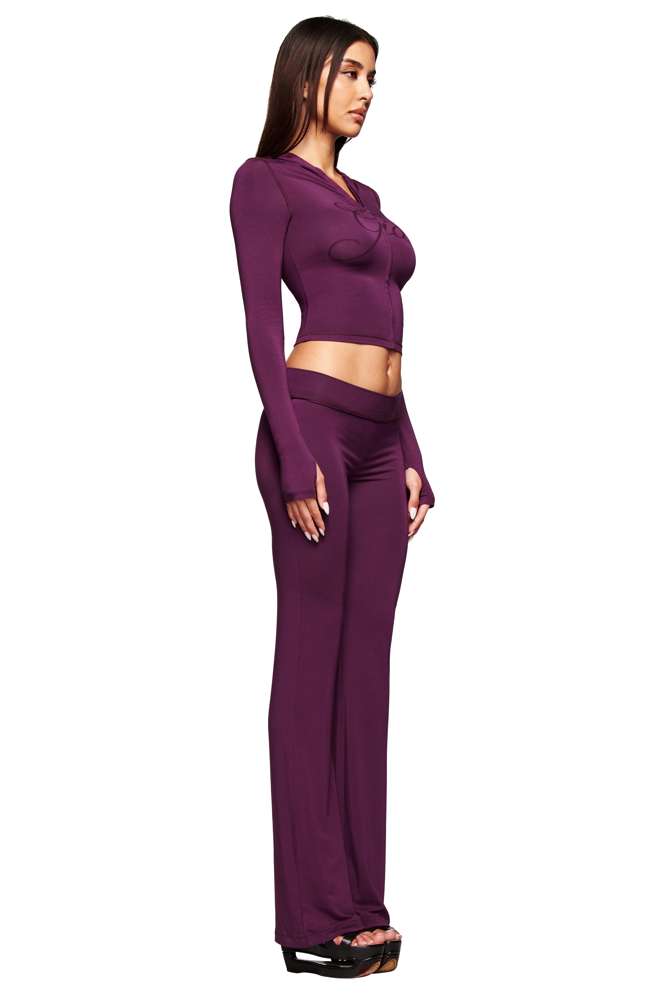 A woman stands in profile against a white background, wearing the I.AM.GIA BLARE TRACKPANT - PLUM with a fitted, long-sleeve, matching crop top and black open-toe heels.