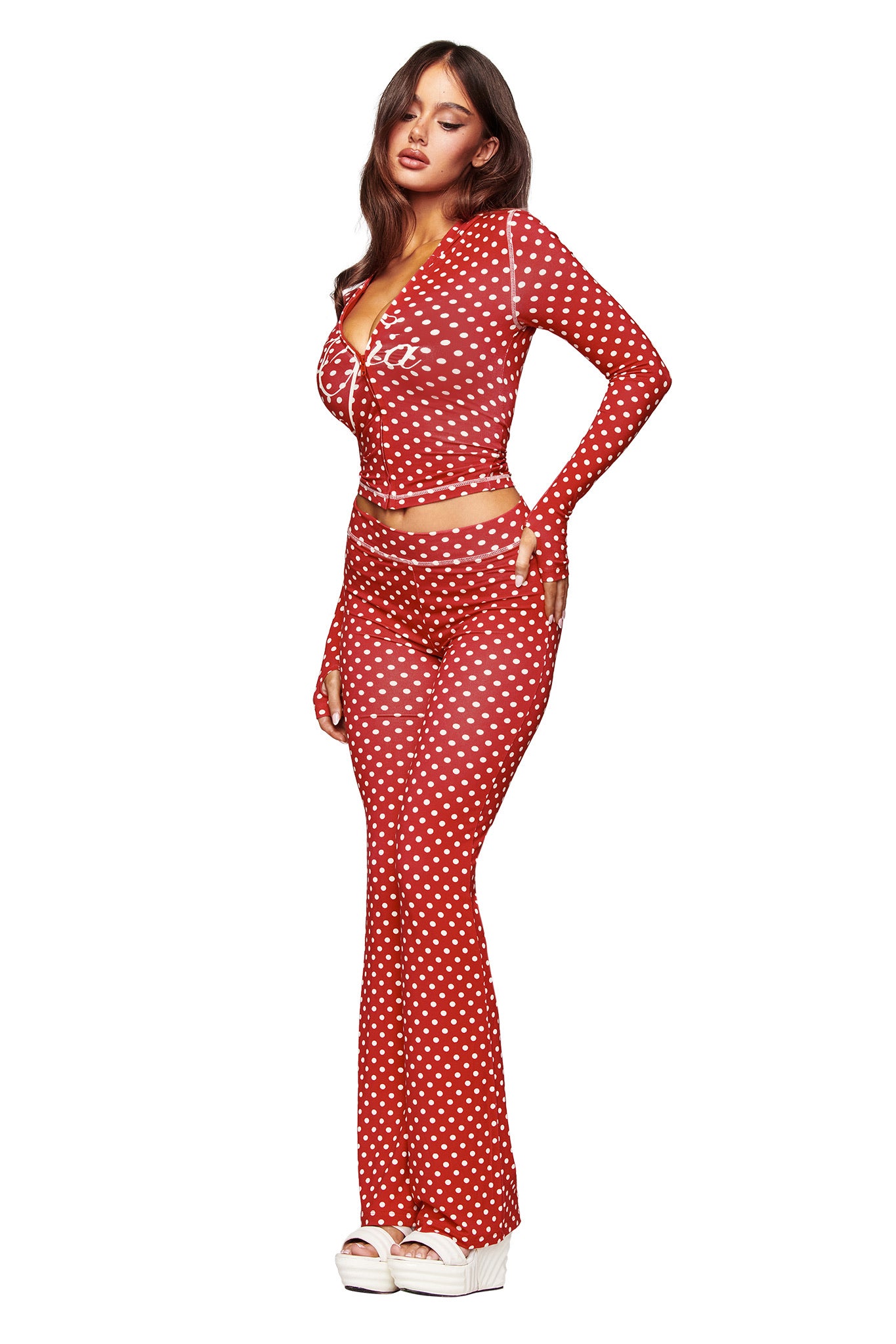 A woman poses against a white background in the I.AM.GIA BLARE TRACKPANT - RED POLKA DOT set, featuring a red polka dot long-sleeve top and trackpants crafted from soft bamboo fabric, styled with white platform sandals.