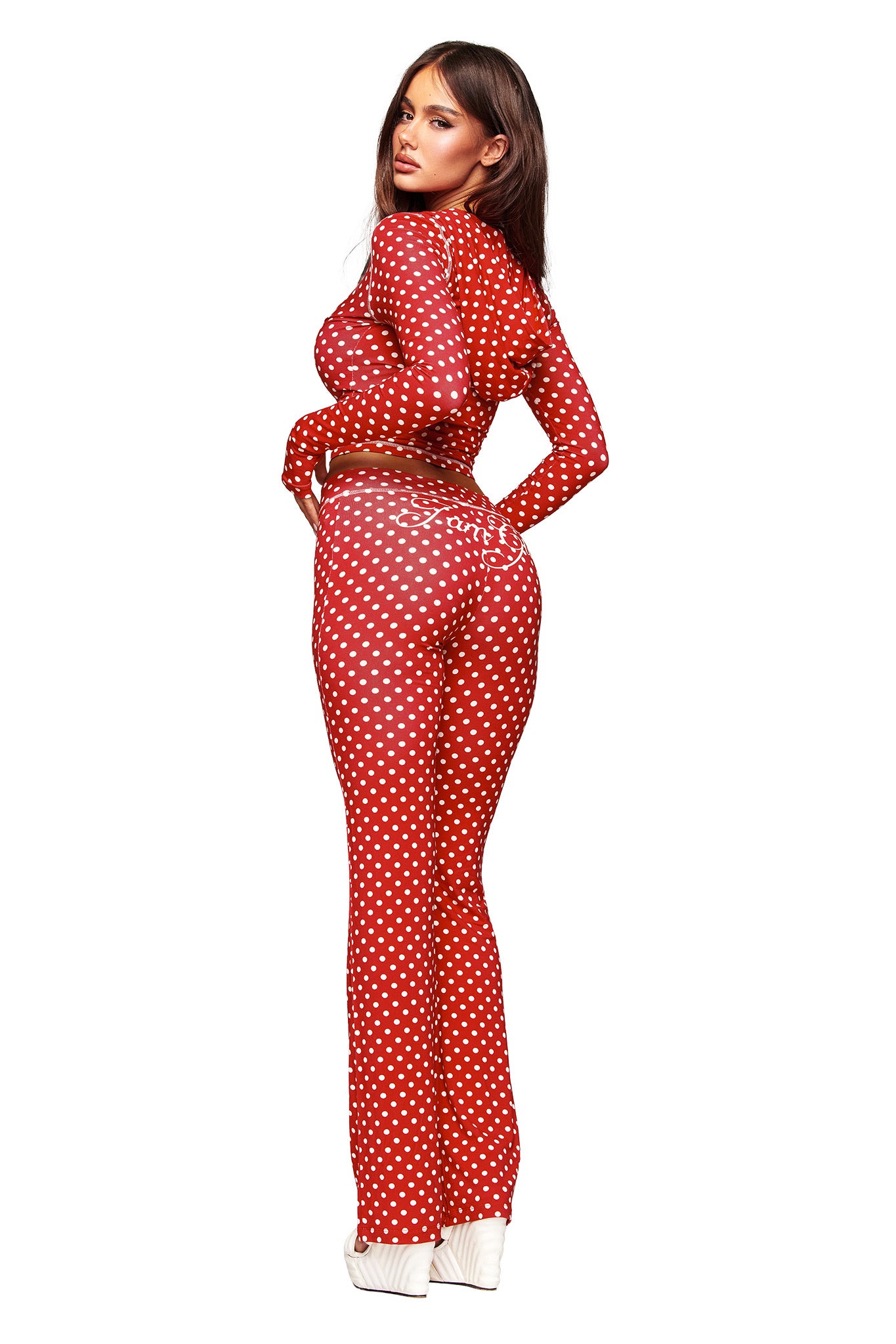 A woman poses in the I.AM.GIA BLARE HOODIE - RED POLKA DOT, a fitted long-sleeve jumpsuit. She looks over her shoulder, wearing white platform shoes, with long brown hair against a plain white background.