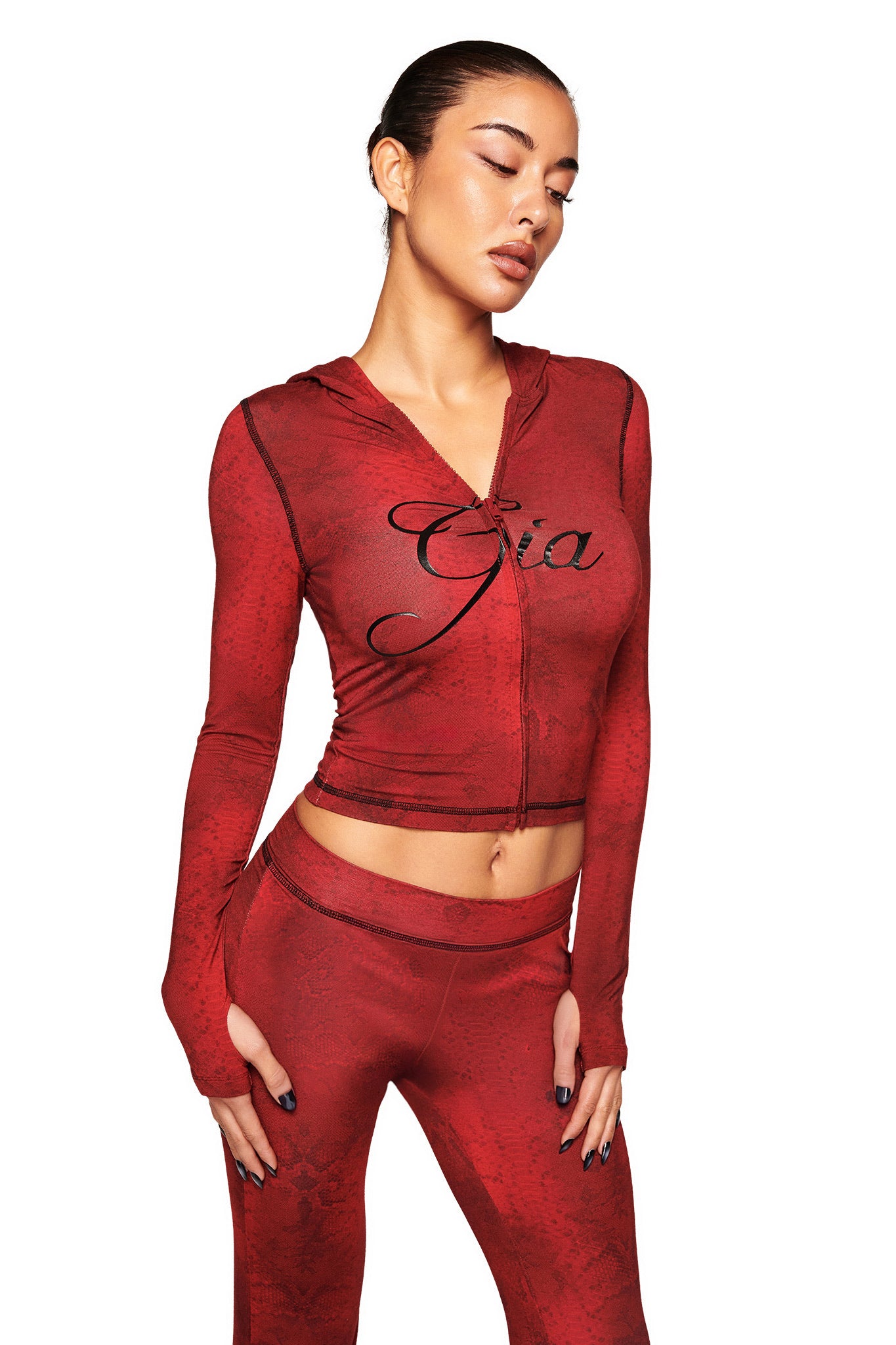 A woman in a fitted red BLARE HOODIE - RED SERPENTINE by I.AM.GIA, featuring “Gia” on the chest and matching pants, stands against a white background with her eyes closed, looking down.