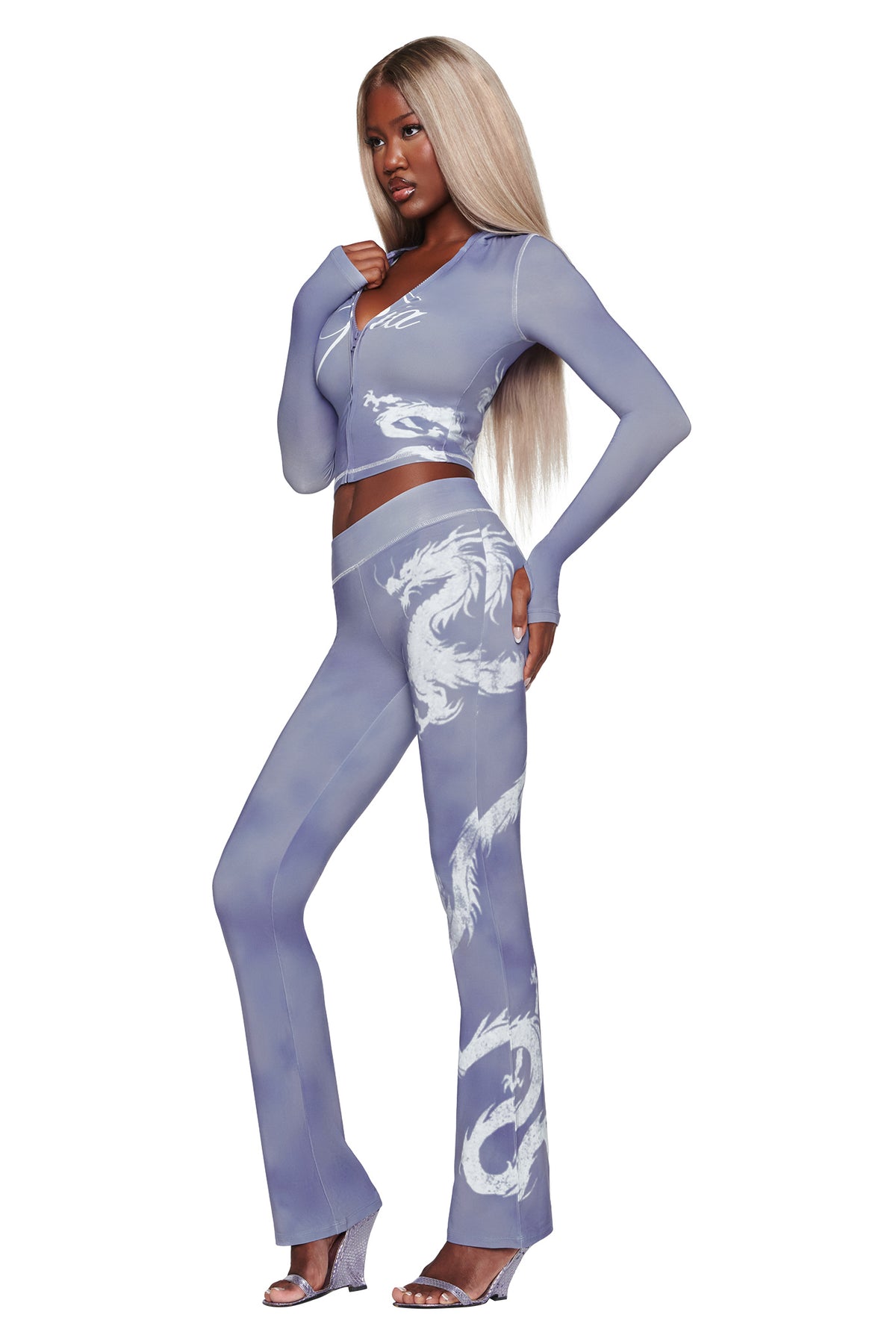 A woman with long blonde hair wears the I.AM.GIA BLARE HOODIE in Steel Blue Dragon, a sheer, fitted light purple hoodie featuring white dragon designs, paired with matching flared pants and clear high-heeled shoes, standing against a white background.