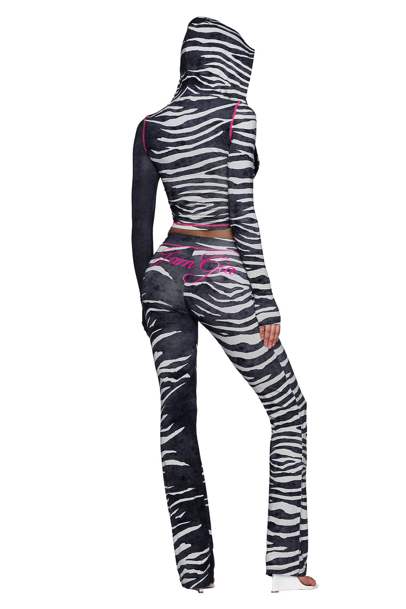 A person wearing the I.AM.GIA BLARE TRACKPANT - BLACK WILD SIDE, a zebra-print trackpant with a mid-rise waist and pink trim, along with a matching hooded jacket featuring “Team Gia” in pink on the back, stands with their back to the camera.