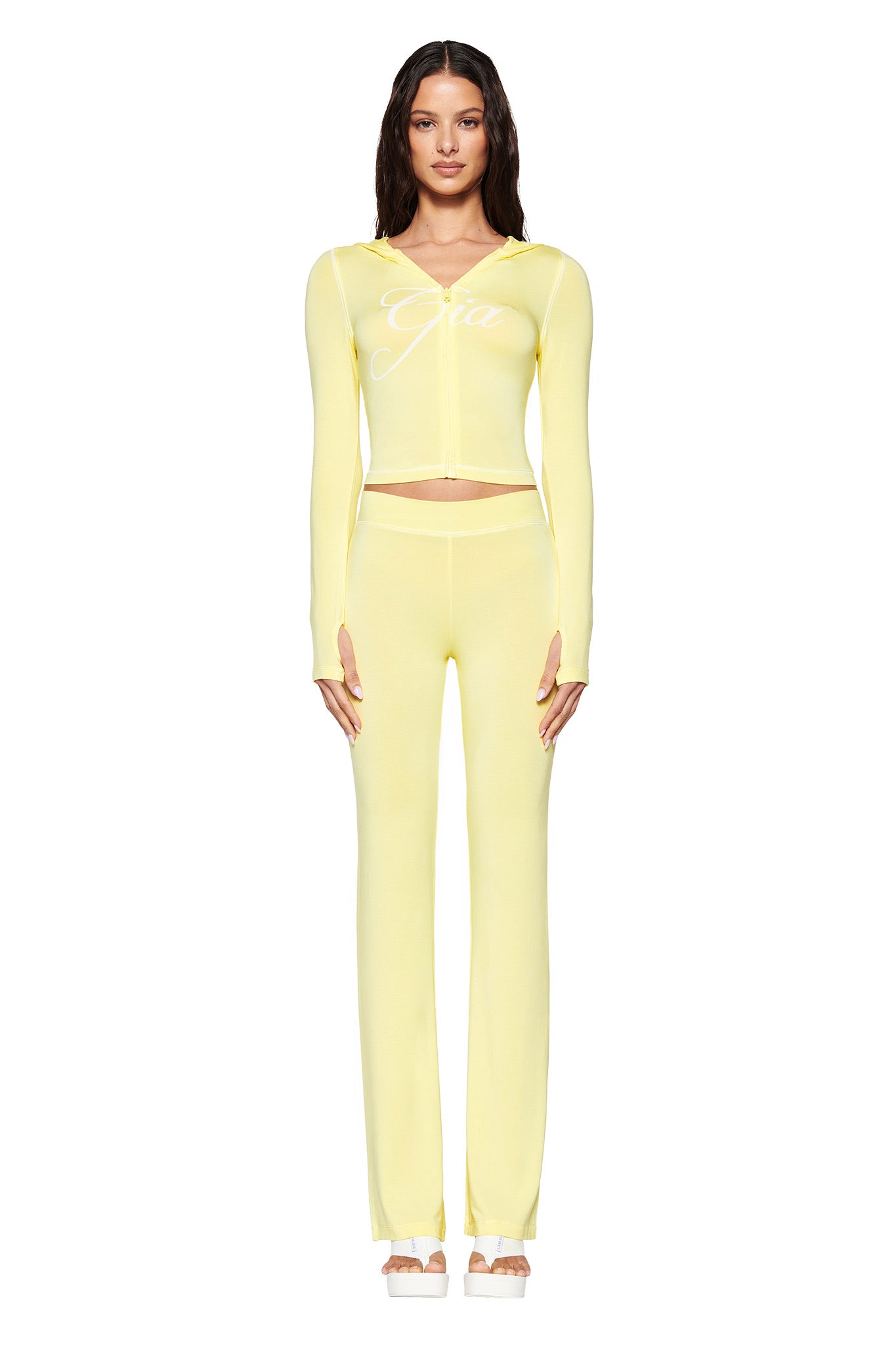 A person stands facing forward in a pale yellow two-piece set from I.AM.GIA, featuring the BLARE HOODIE with "Gia" printed on it, thumb-hole cut-outs, and matching high-waisted pants. The background is white.
