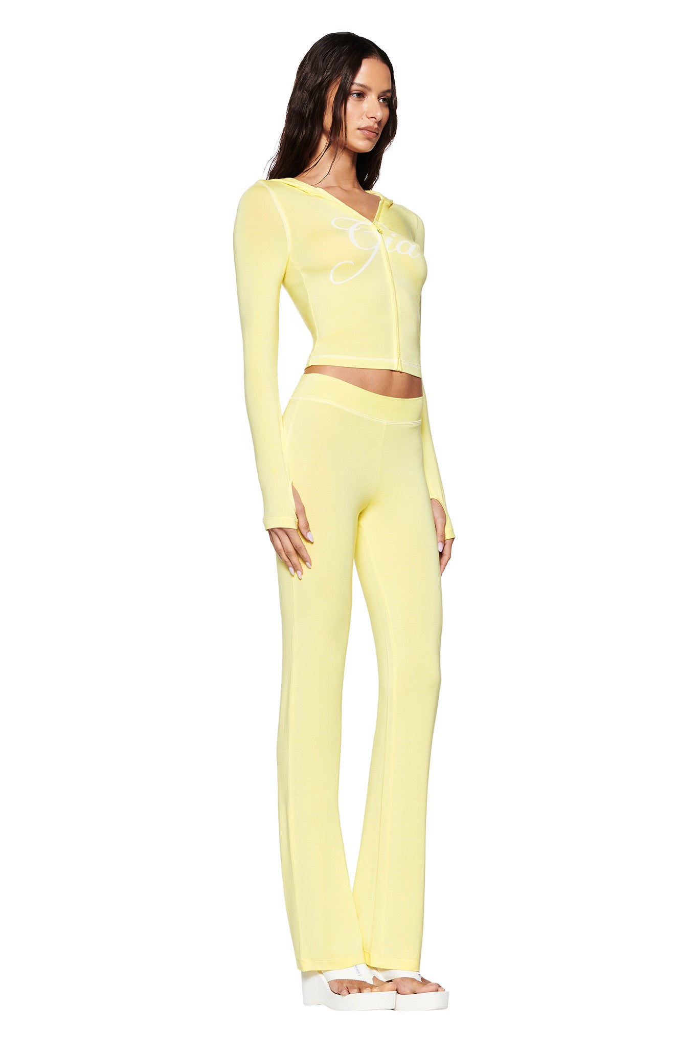 A woman wears the pale yellow BLARE HOODIE by I.AM.GIA, featuring long sleeves with thumb-hole cut-outs and matching flare pants. Made from stretchy bamboo fabric, it sports a bold white cursive print on the chest. She stands sideways, her head turned slightly left, completing her look with platform shoes.