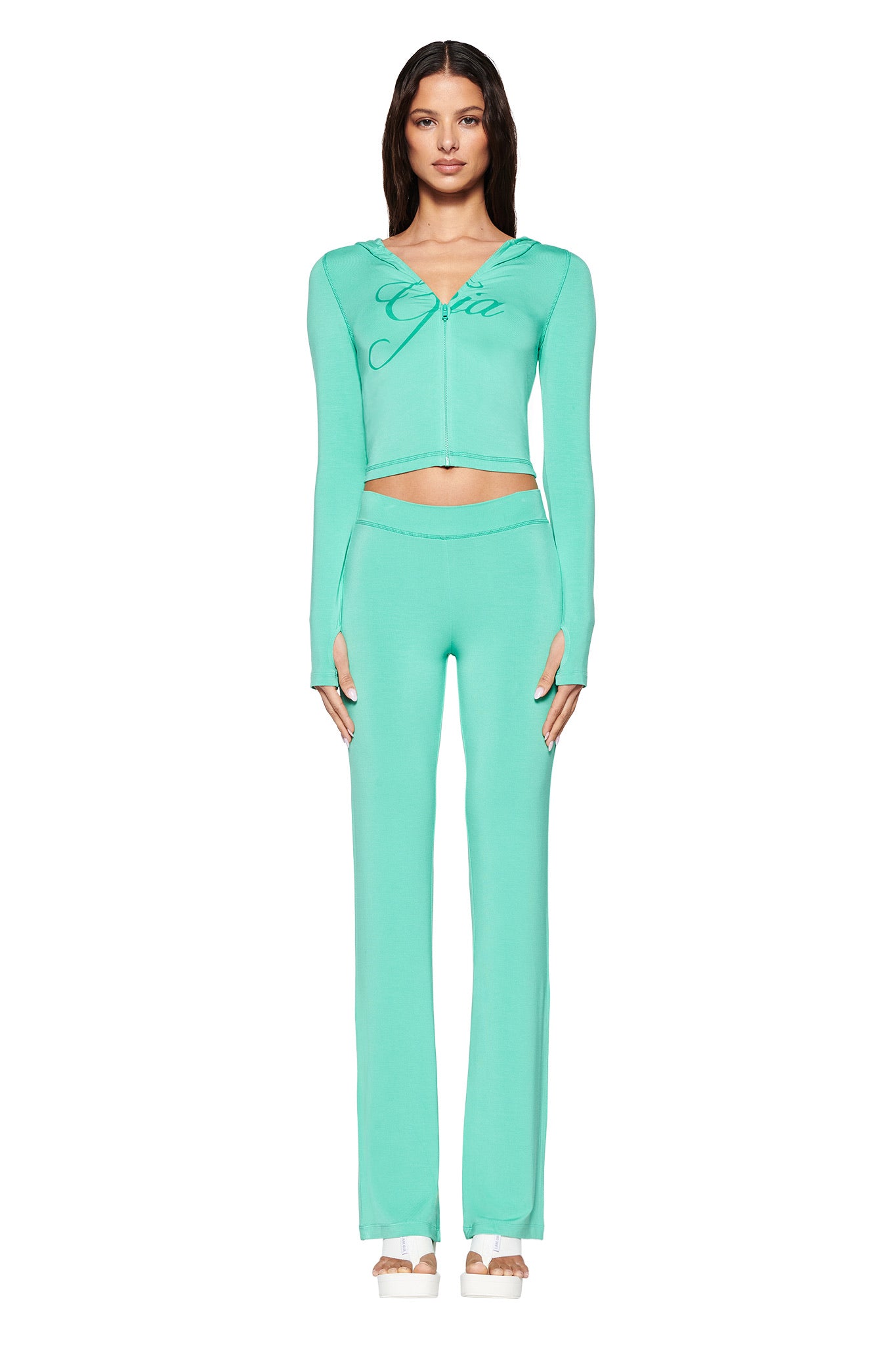 An individual stands against a white background wearing the I.AM.GIA BLARE HOODIE in turquoise teal. The long-sleeve top with thumb holes and script writing creates a bodycon silhouette, paired with matching high-waisted pants and nude platform sandals.