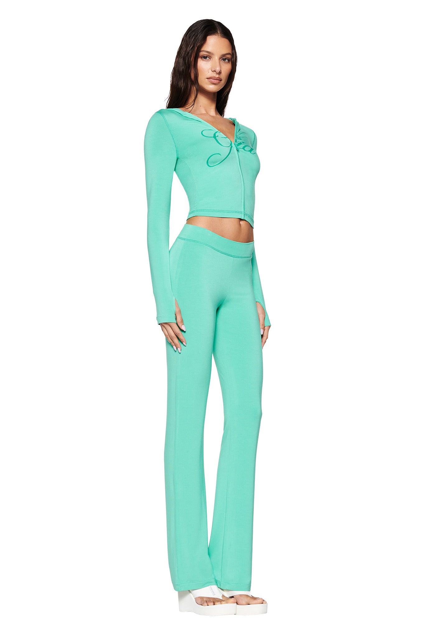 A woman poses in a side stance wearing the I.AM.GIA BLARE HOODIE in turquoise teal with matching flared pants, showcasing a bow detail at the neckline. Her look is completed with white platform shoes against a plain white background.