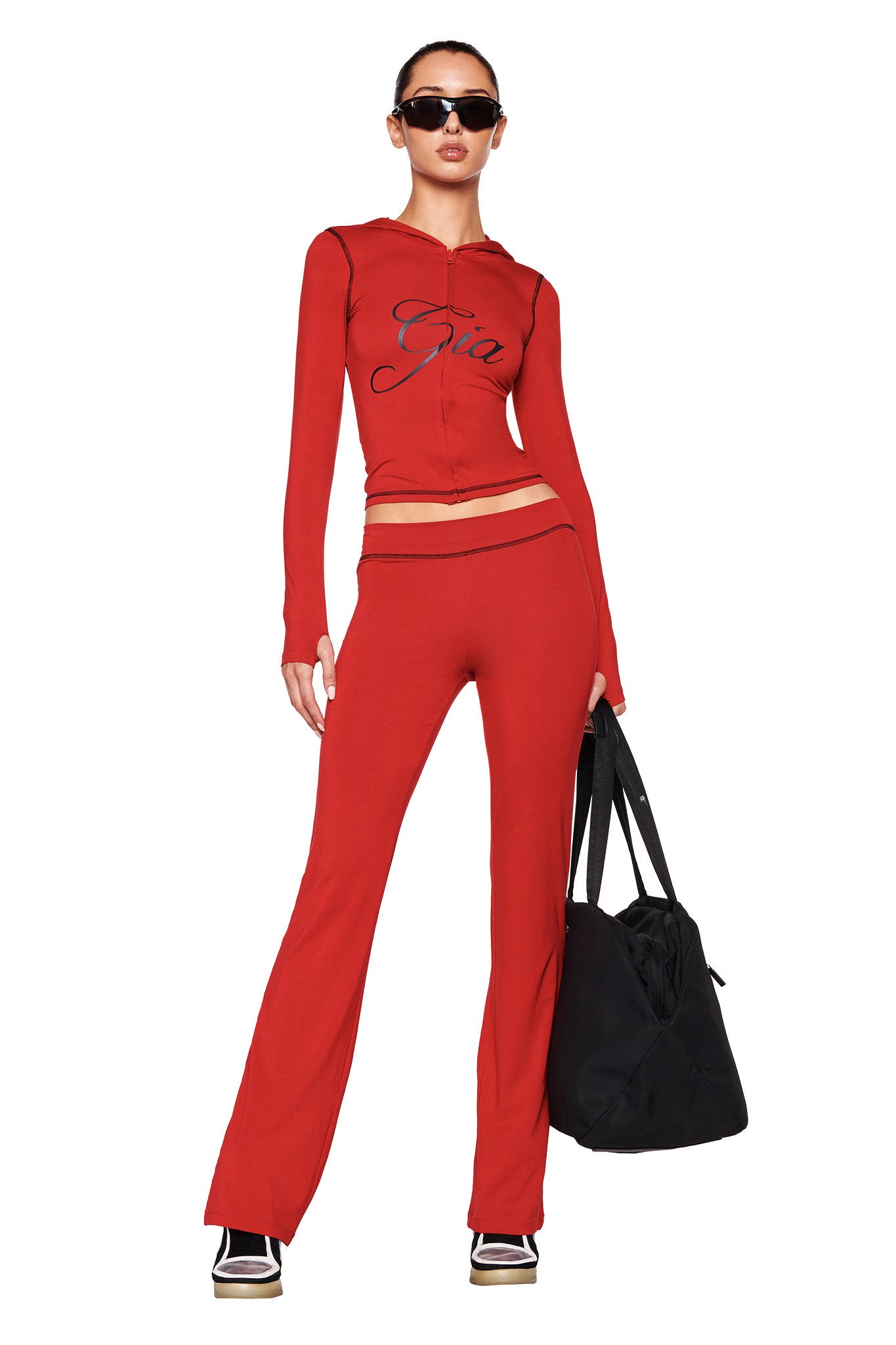 A person wearing a red, bodycon silhouette long-sleeve top with the word "Gia" printed on it and matching I.AM.GIA BLARE TRACKPANT - RED made from stretchy bamboo fabric. They are also wearing black and white sneakers, sunglasses, and holding a large black bag. The person has their hair pulled back and appears confident.
