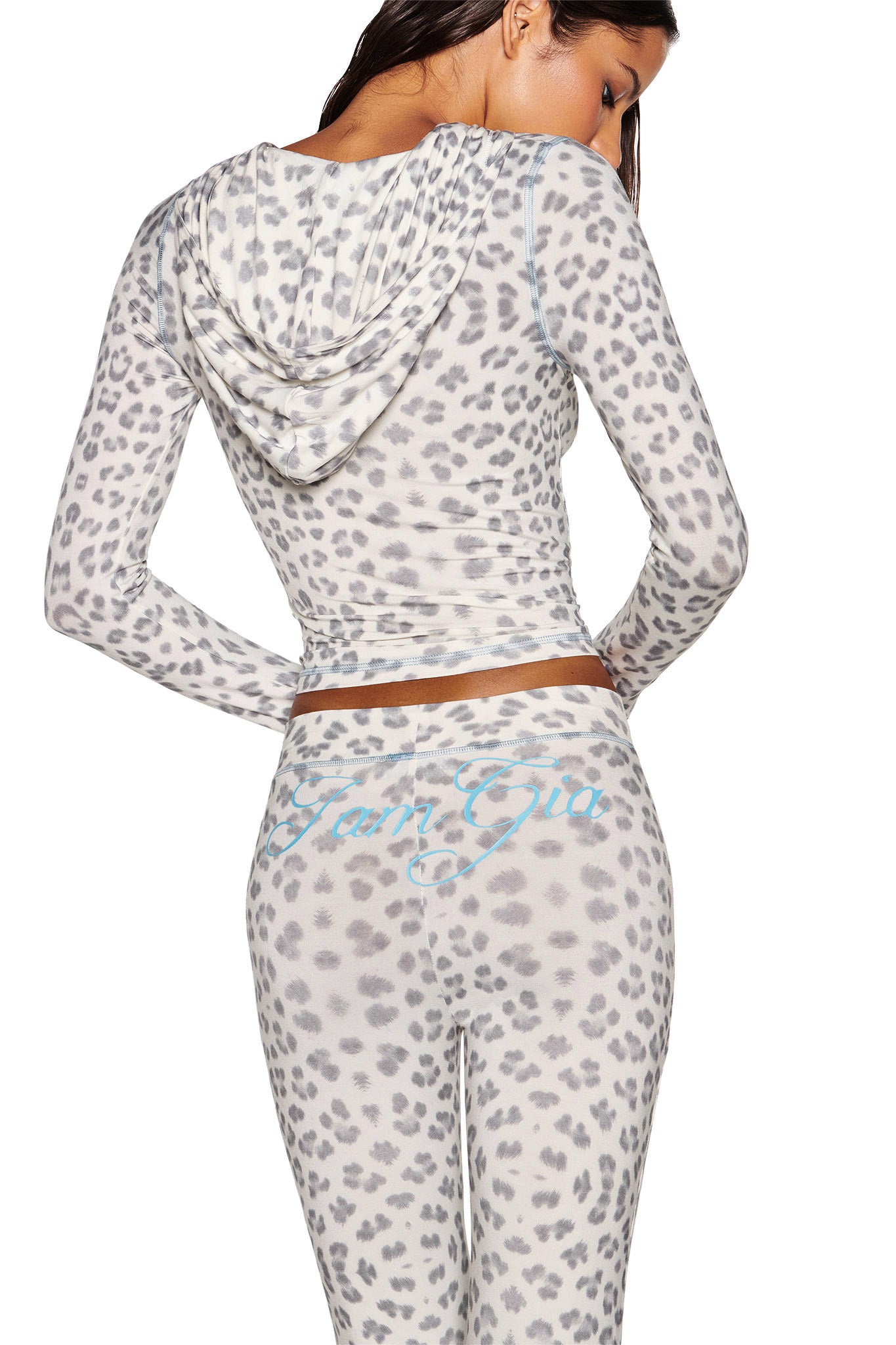 A woman, facing away from the camera, models a bodycon outfit in a snow leopard print. The I.AM.GIA Blare Hoodie - Snow Leopard: Winter Wild Card features long sleeves, paired with leggings adorned with light blue cursive writing. The setting is against a sharp white background.