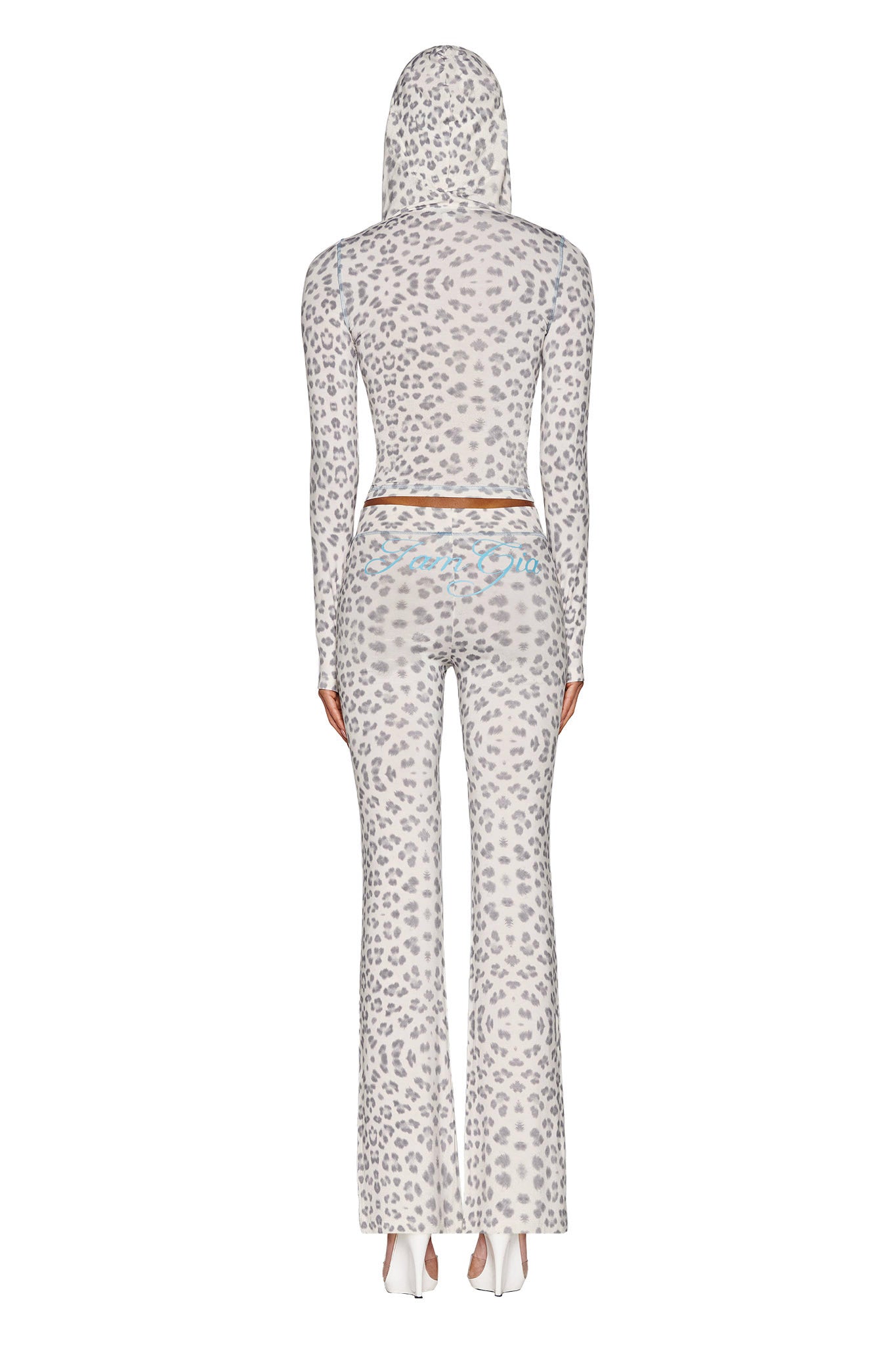 A person dressed in the I.AM.GIA BLARE HOODIE - SNOW LEOPARD from the WINTER WILD CARD collection, paired with matching flared pants, is standing with their back to the camera. The bodycon fit outfit showcases a subtle blue design on the waistband and is perfectly complemented by sleek white heels.