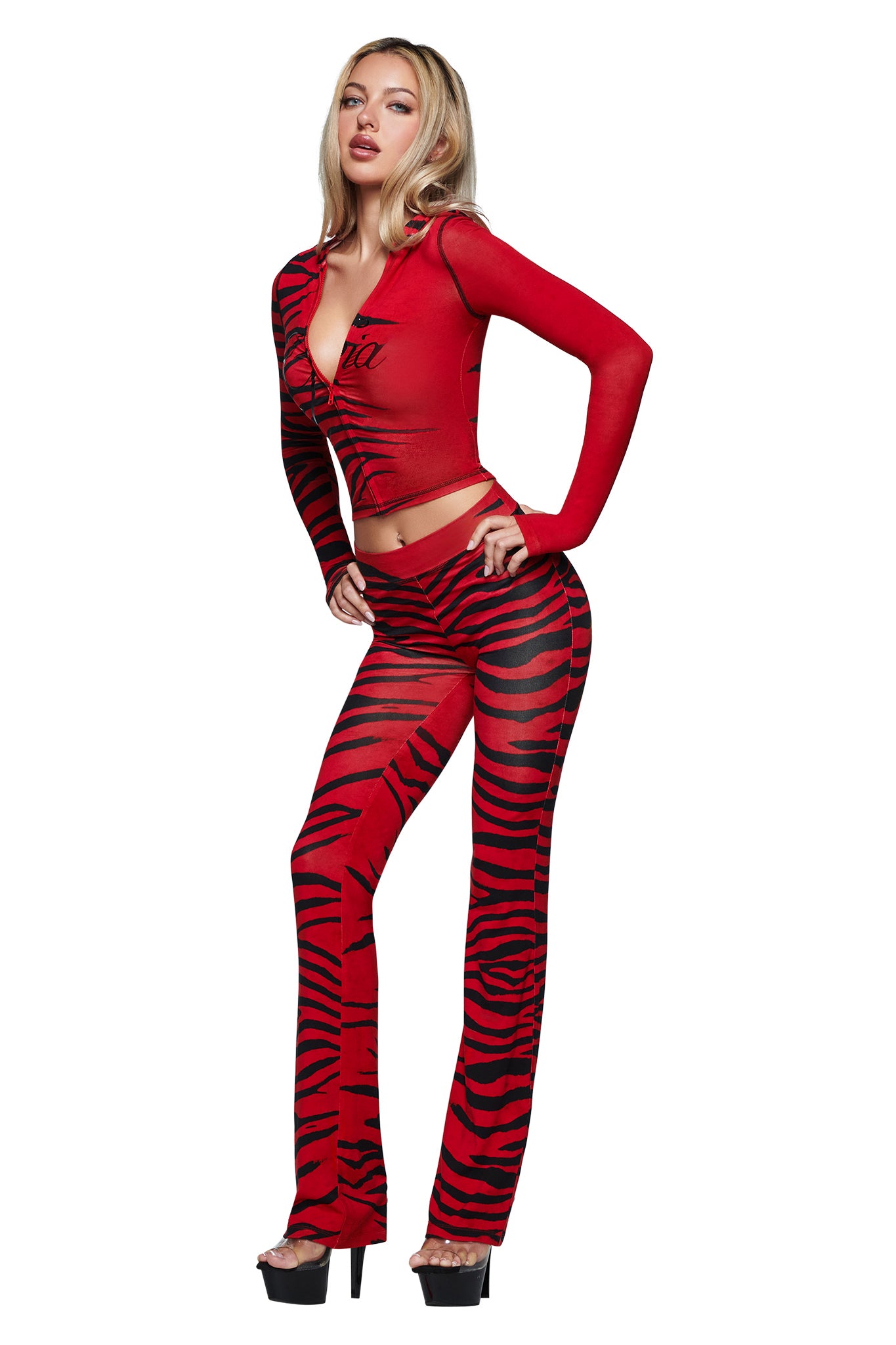 A woman poses in the BLARE TRACKPANT - RED WILD SIDE by I.AM.GIA with a matching long-sleeve crop top featuring the brand’s logo, paired with black high heels against a white background.