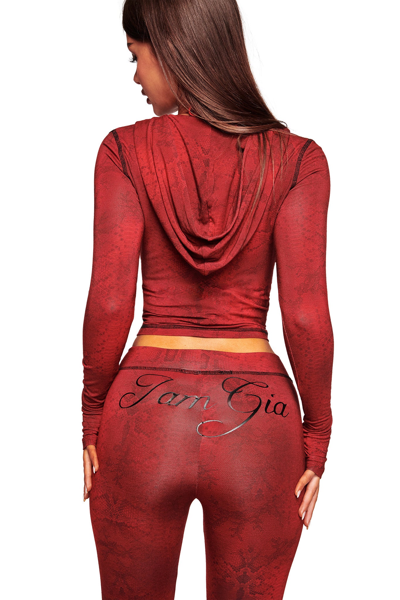 A woman stands with her back to the camera, wearing the fitted BLARE HOODIE - RED SERPENTINE by I.AM.GIA. Her long brown hair falls over the hoodie's red fabric; "I.AM.GIA" is scripted on her leggings. White background.