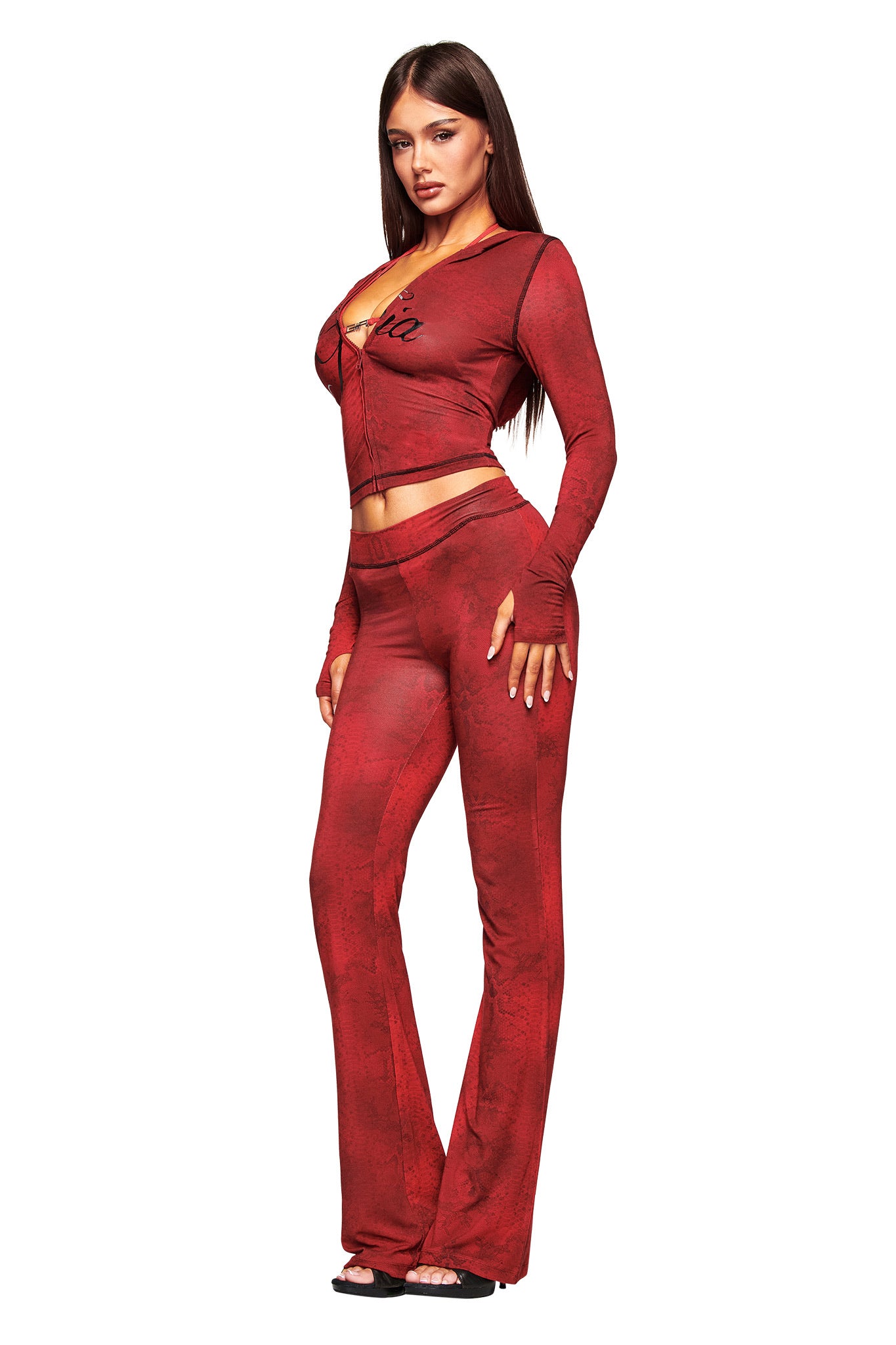 A woman with long brown hair poses with one hand on her hip, wearing the I.AM.GIA BLARE TRACKPANT - RED SERPENTINE featuring a low-rise waist and flared legs, paired with a matching fitted top against a white background.